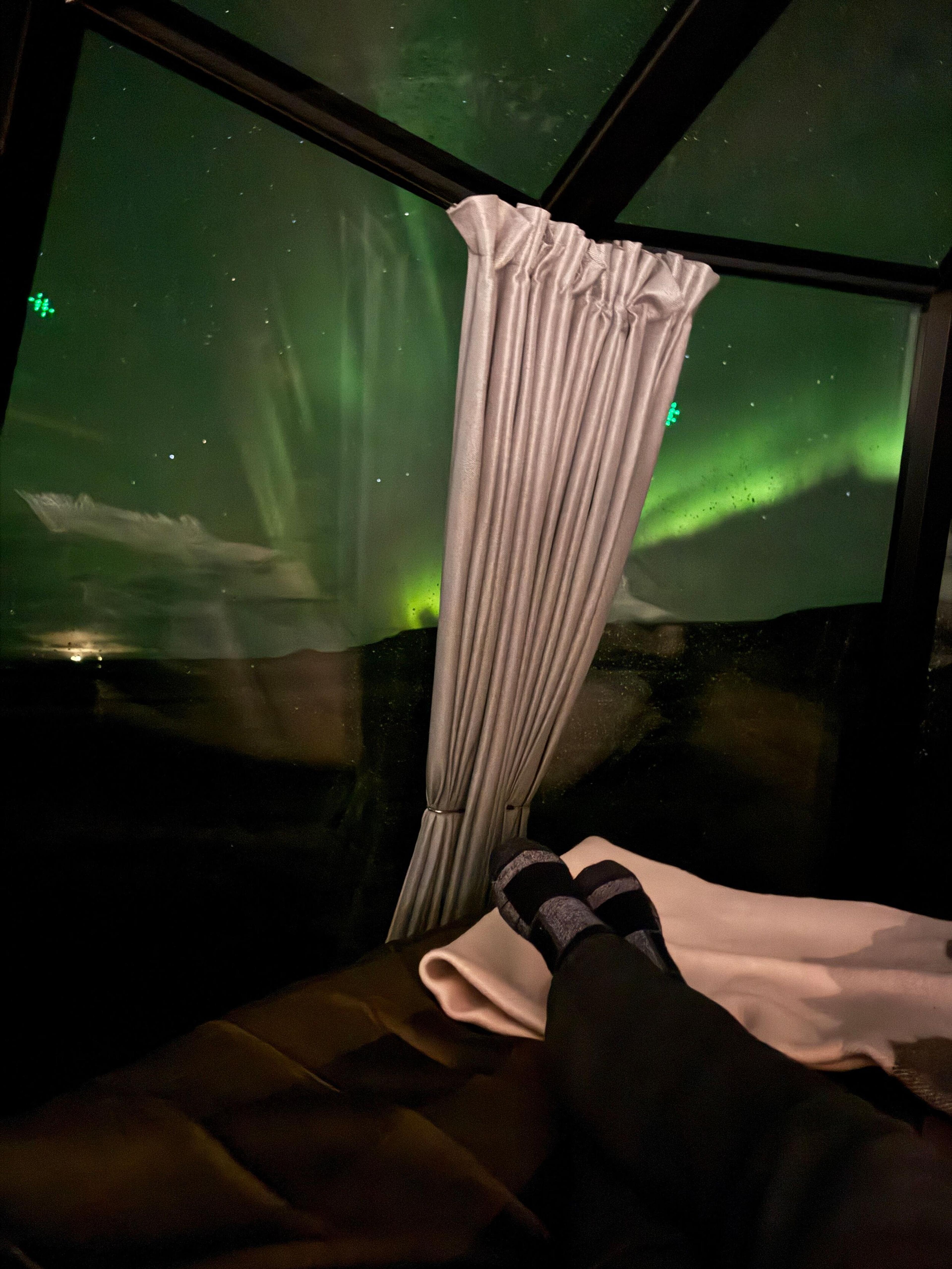 Person relaxing in bed, viewing the Northern Lights through a glass ceiling.