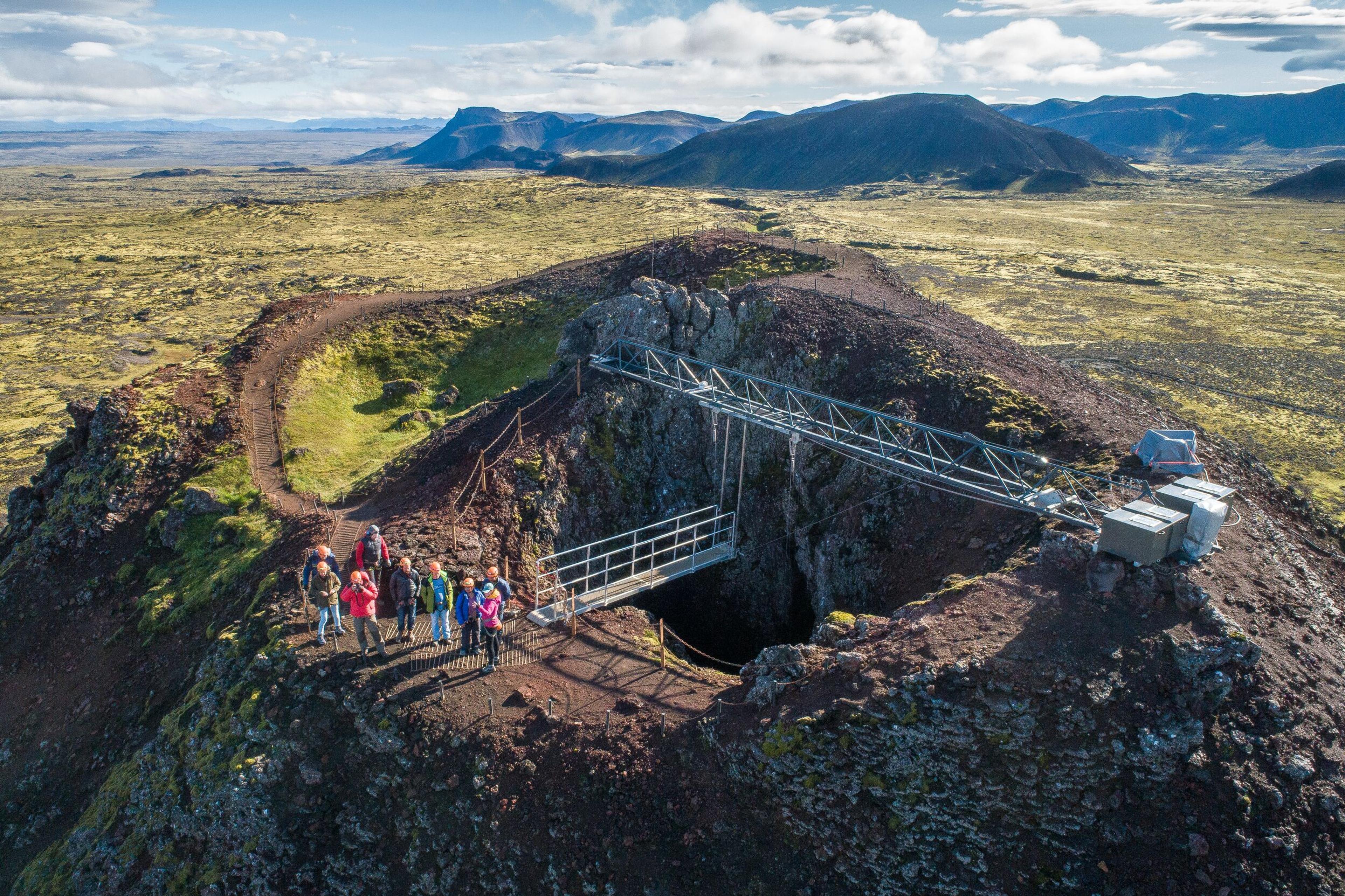 A group of hikers stands at the rim of a volcanic crater equipped with an elevator system, surrounded by a vast, mossy landscape.
