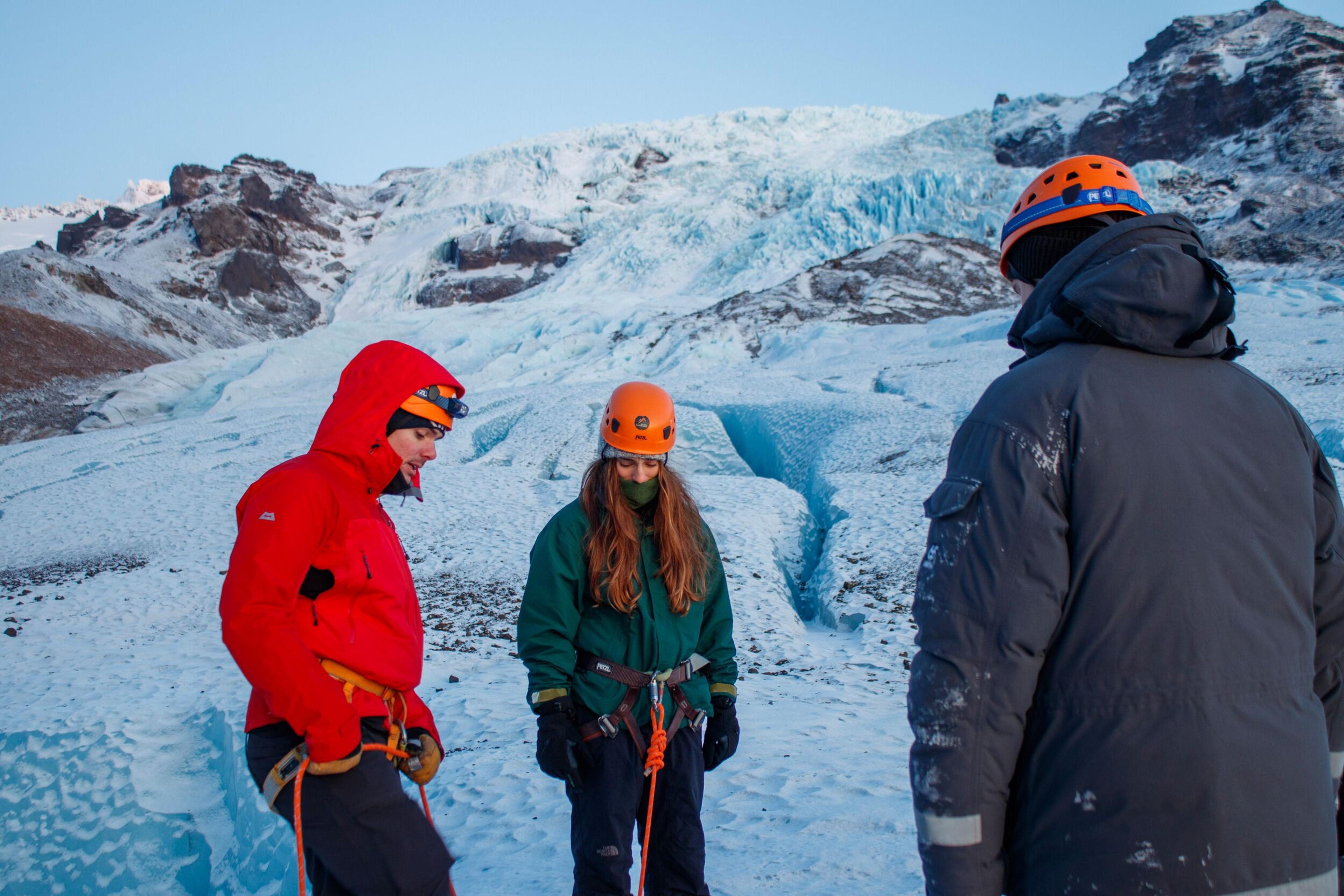 Three people in winter climbing gear stand on a snow-covered glacier with a large ice formation and mountains behind them.