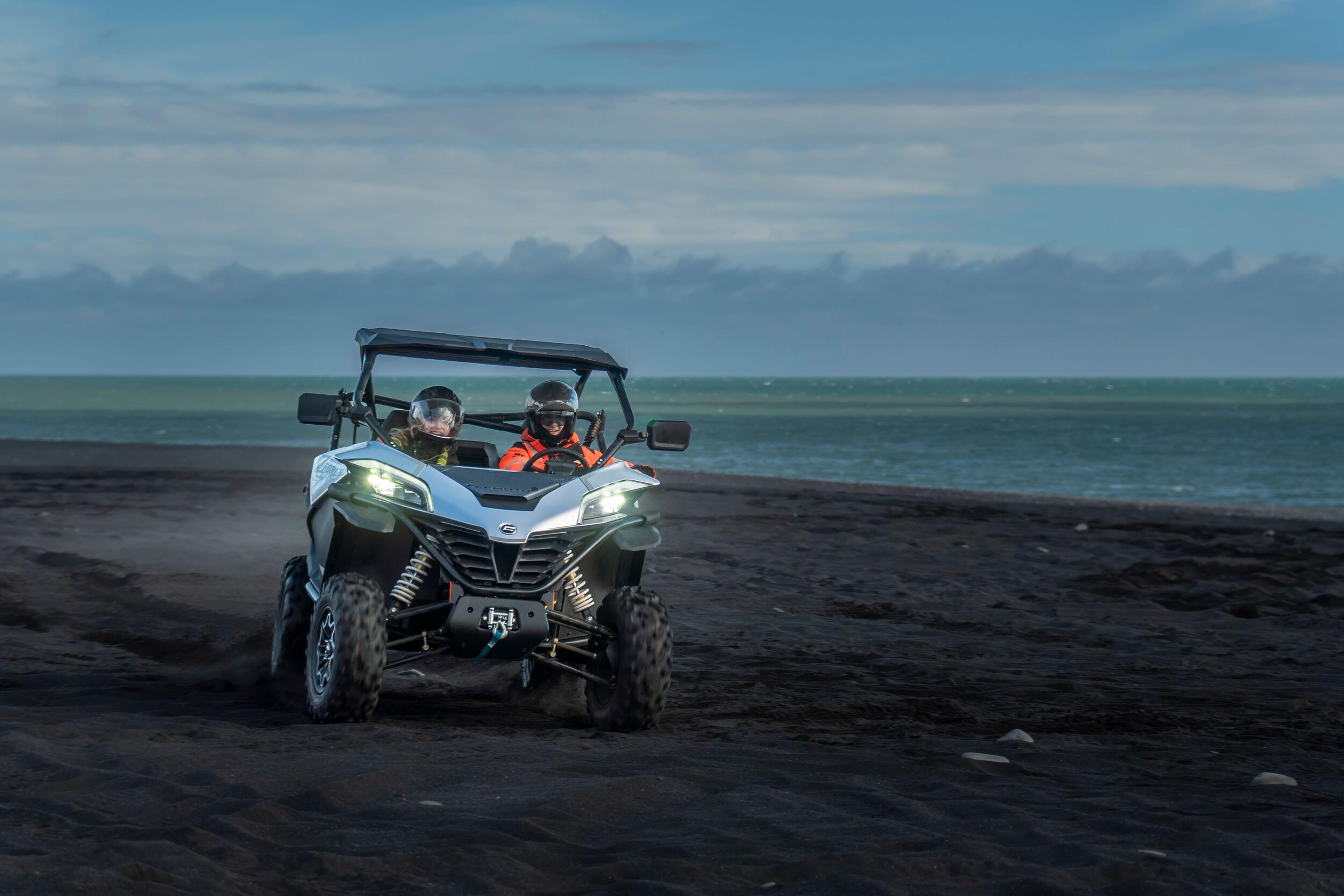 A side-by-side all-terrain vehicle (ATV) with two riders wearing helmets and high-visibility clothing is on a dark sandy beach, with the ocean in the background and a mountain range on the horizon under a twilight sky.