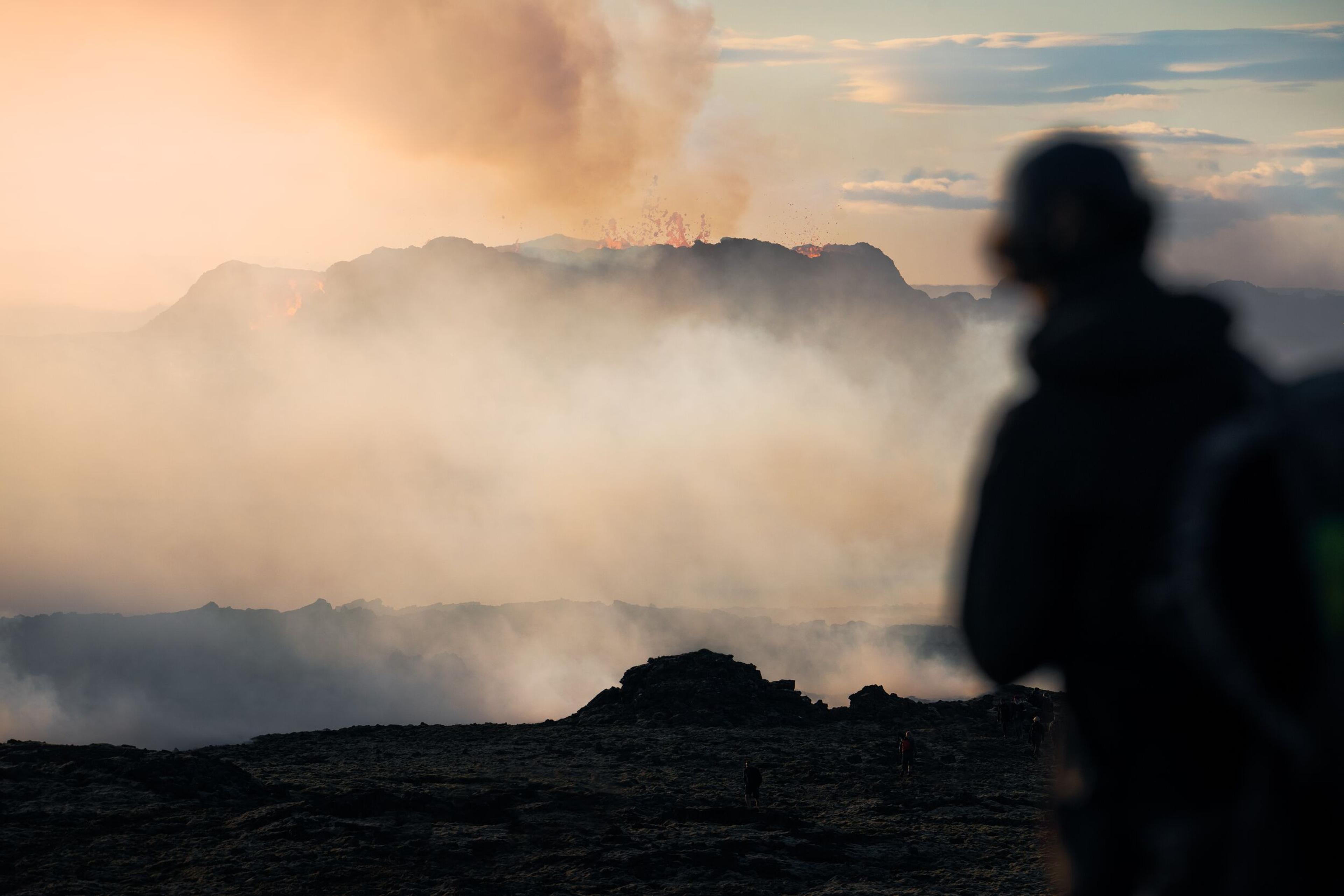 Silhouetted figure against fiery volcanic eruption, with intense blue smoke and orange lava flames.