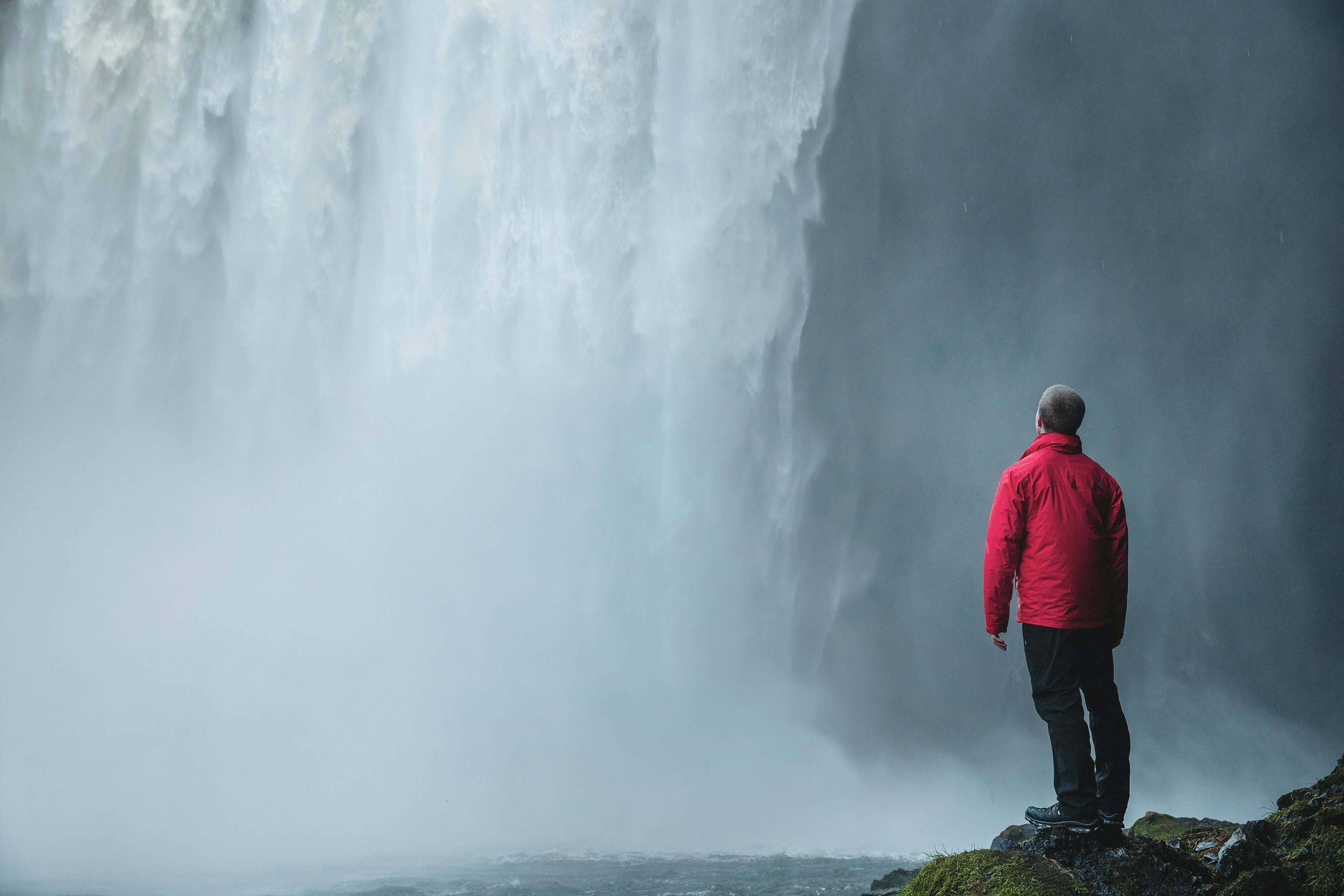 A man in a red jacket stands close to the powerful cascade of Seljalandsfoss waterfall, gazing up at the misty, thunderous water.