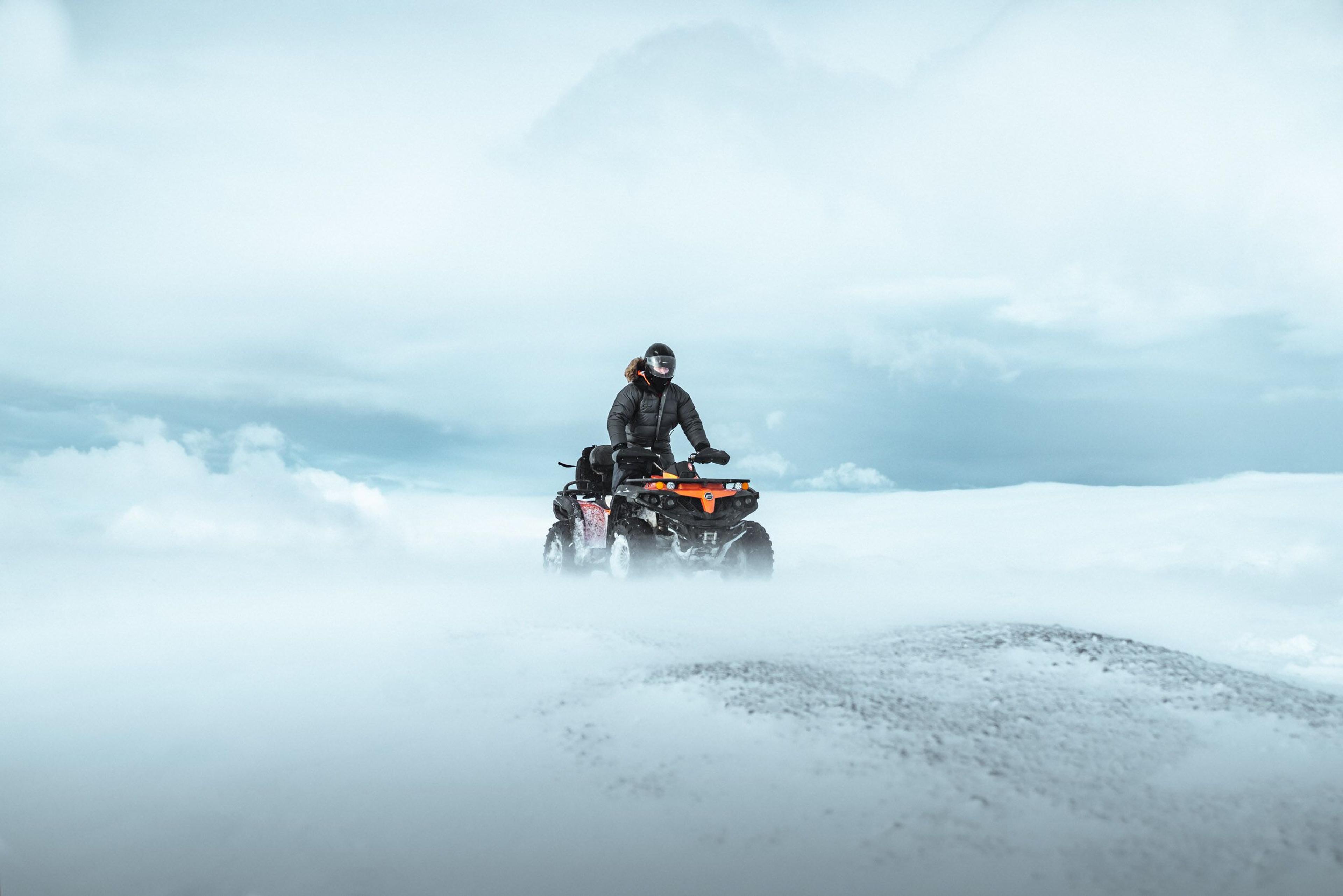 An ATV rider in black winter gear navigates through a vast snowy landscape, surrounded by swirling snow and an overcast sky, creating a striking contrast against the icy terrain.