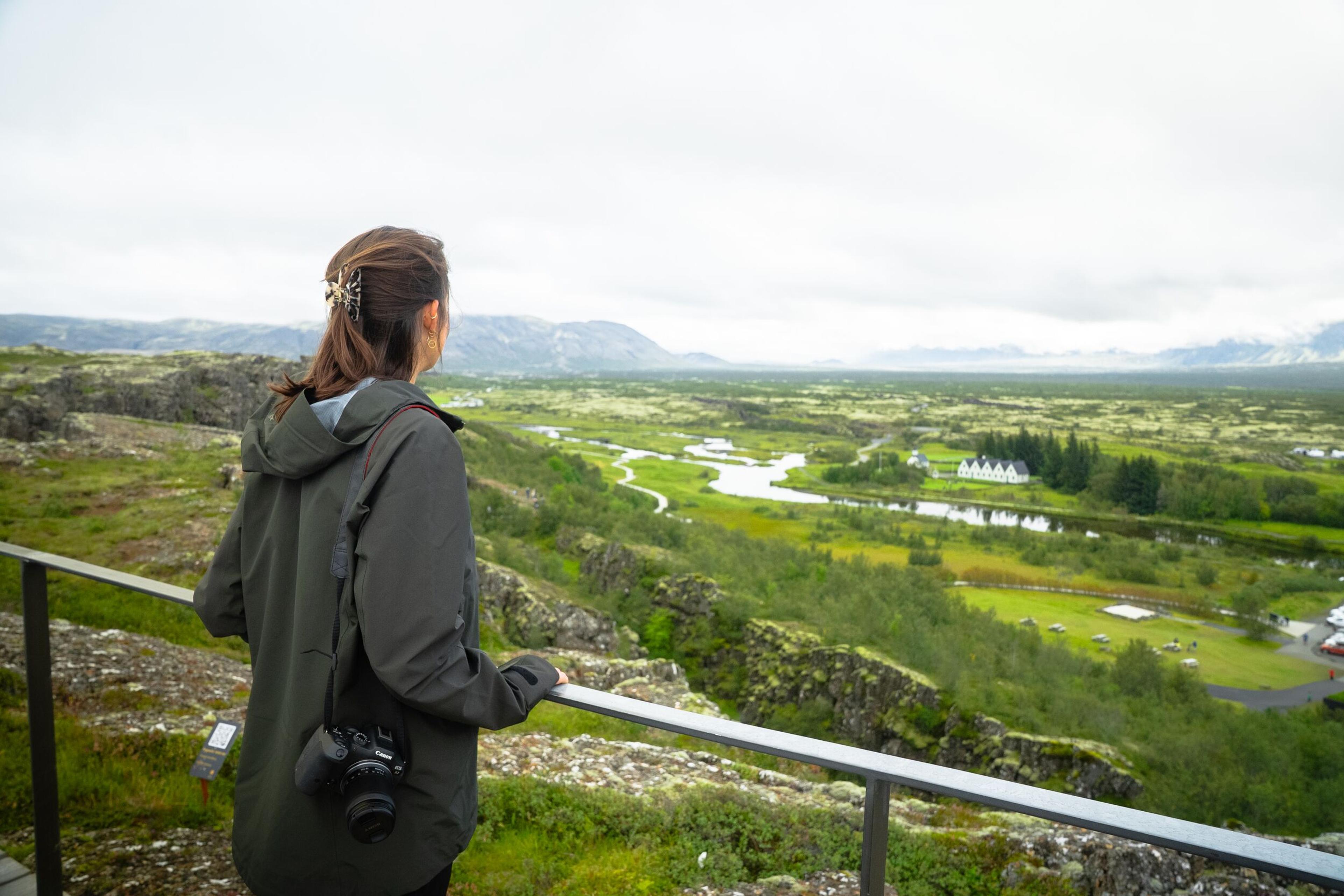 A woman with a camera views a lush green valley with a winding river, buildings, and distant mountains under a cloudy sky.