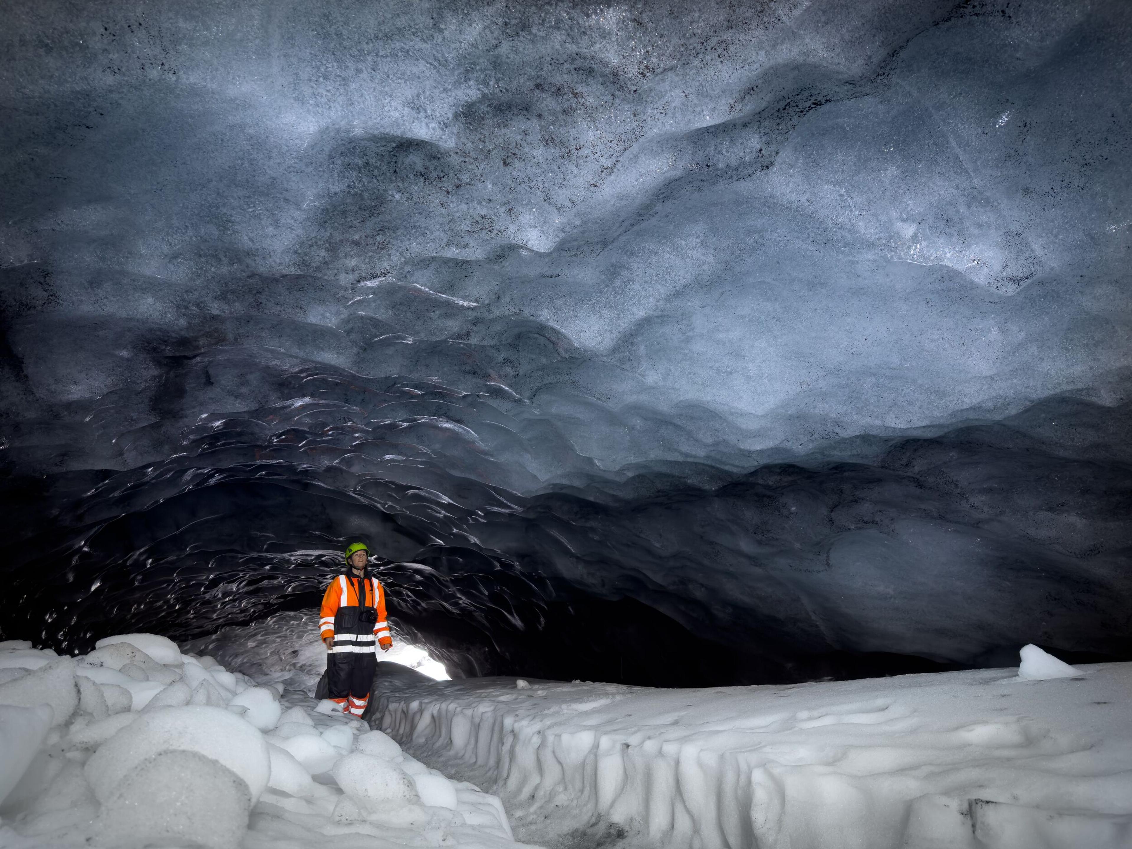 An explorer in safety gear examining the ice ceiling of Askur ice cave, surrounded by snow and ice.