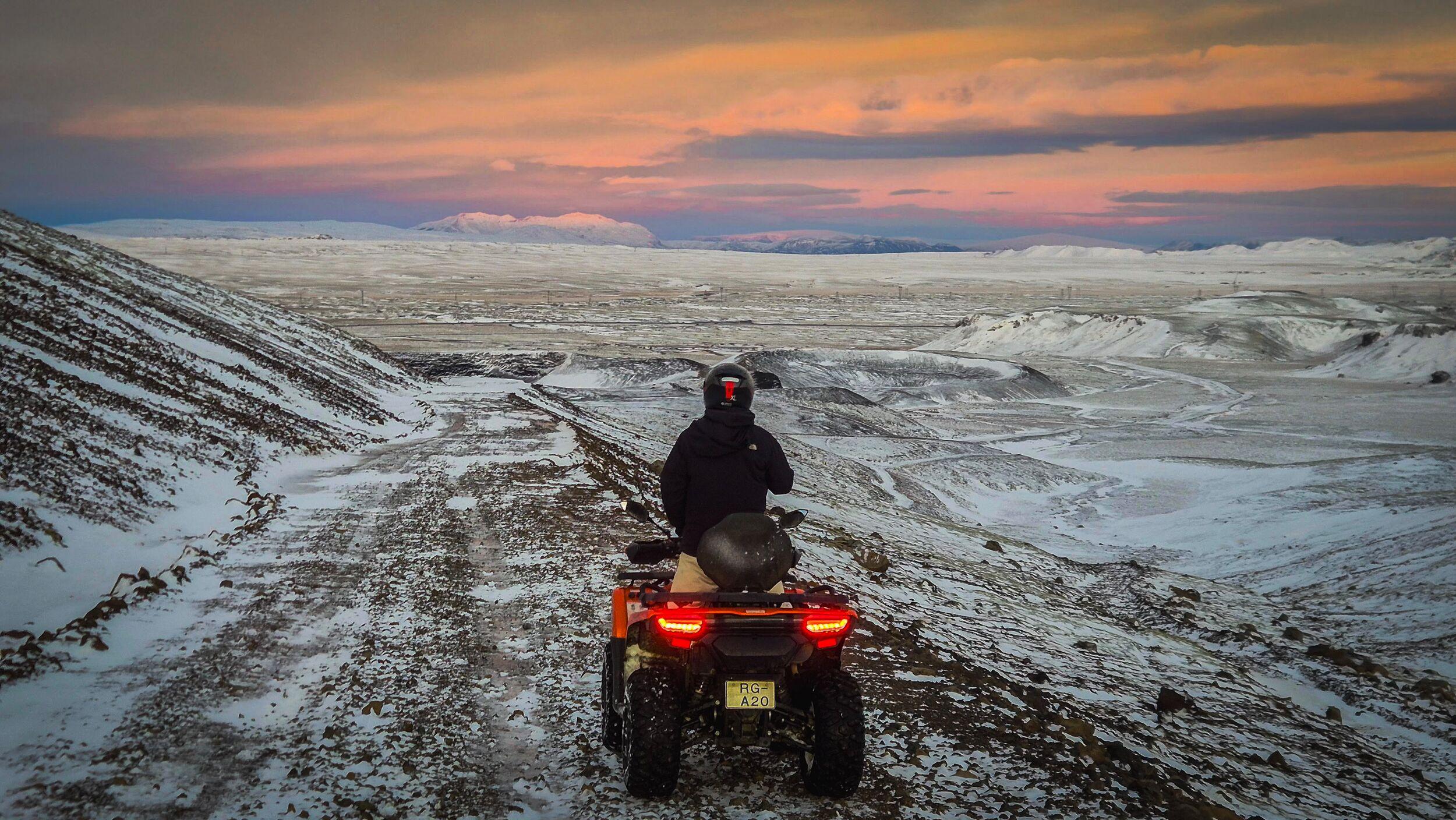 A person on an ATV overlooks a vast, snow-covered winter landscape under a colorful sunset sky.