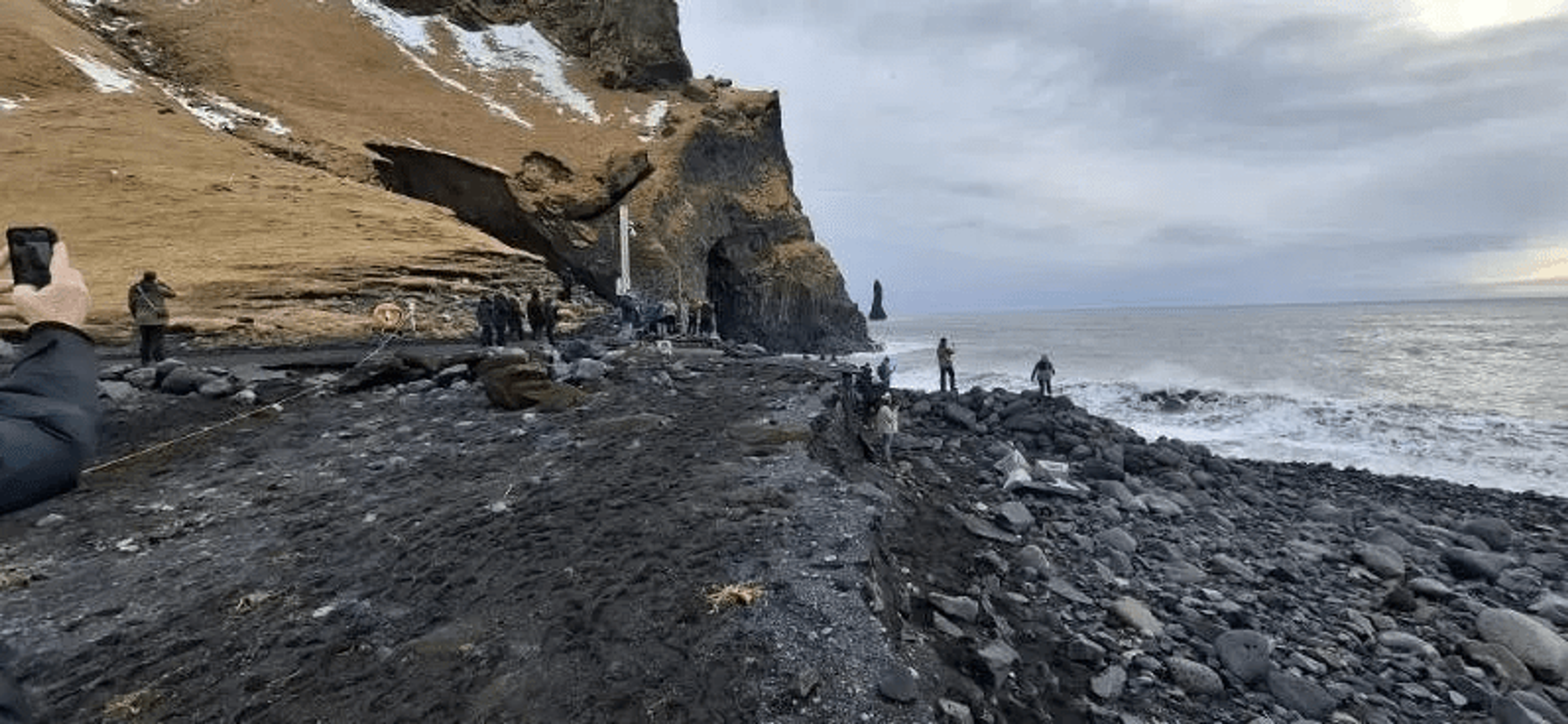 Black rocky beach with a large cliff featuring a sea cave, rough ocean waves, and several people under an overcast sky.