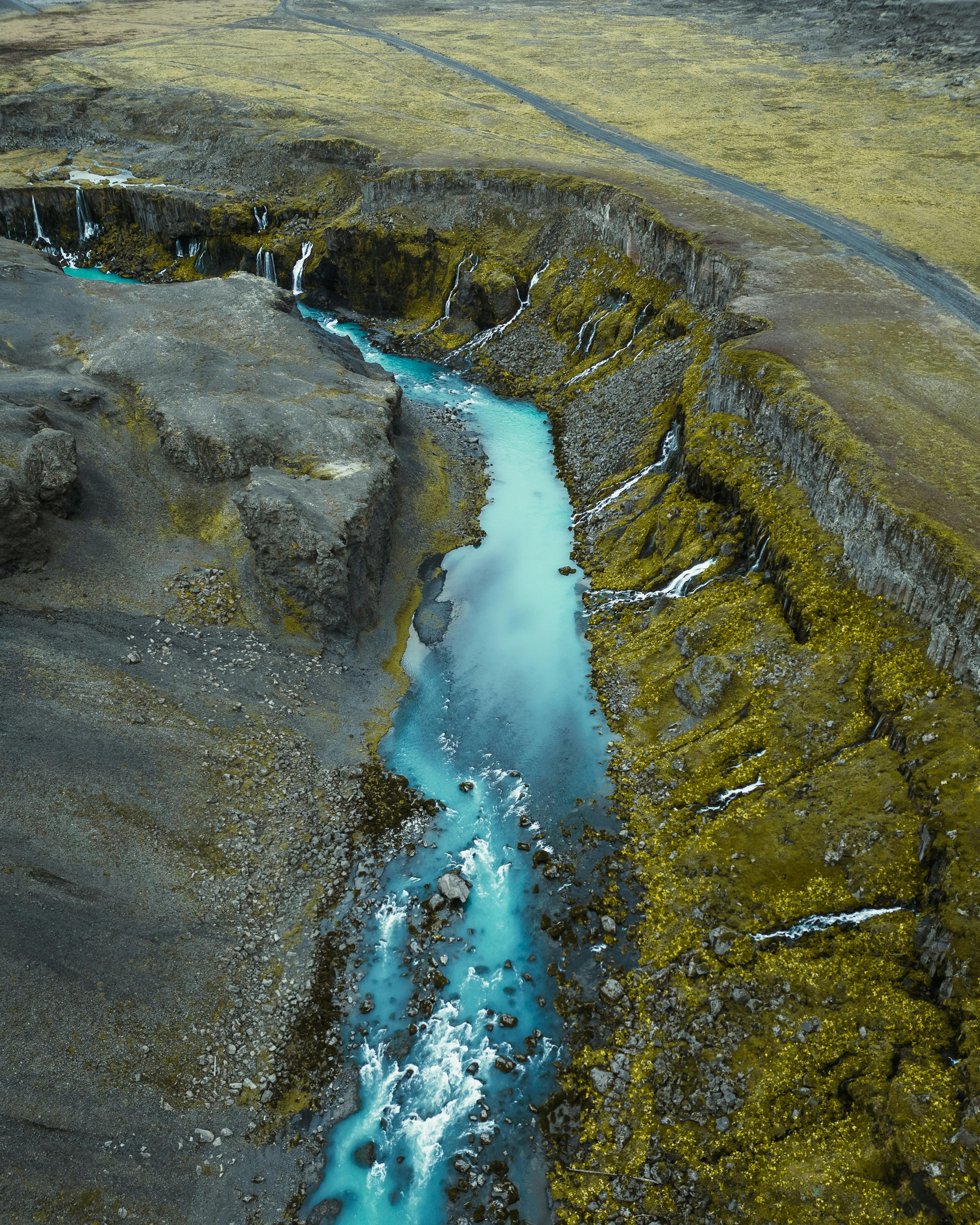 "A vibrant blue river winding through a moss-covered canyon with small cascading waterfalls in Iceland's rugged landscape