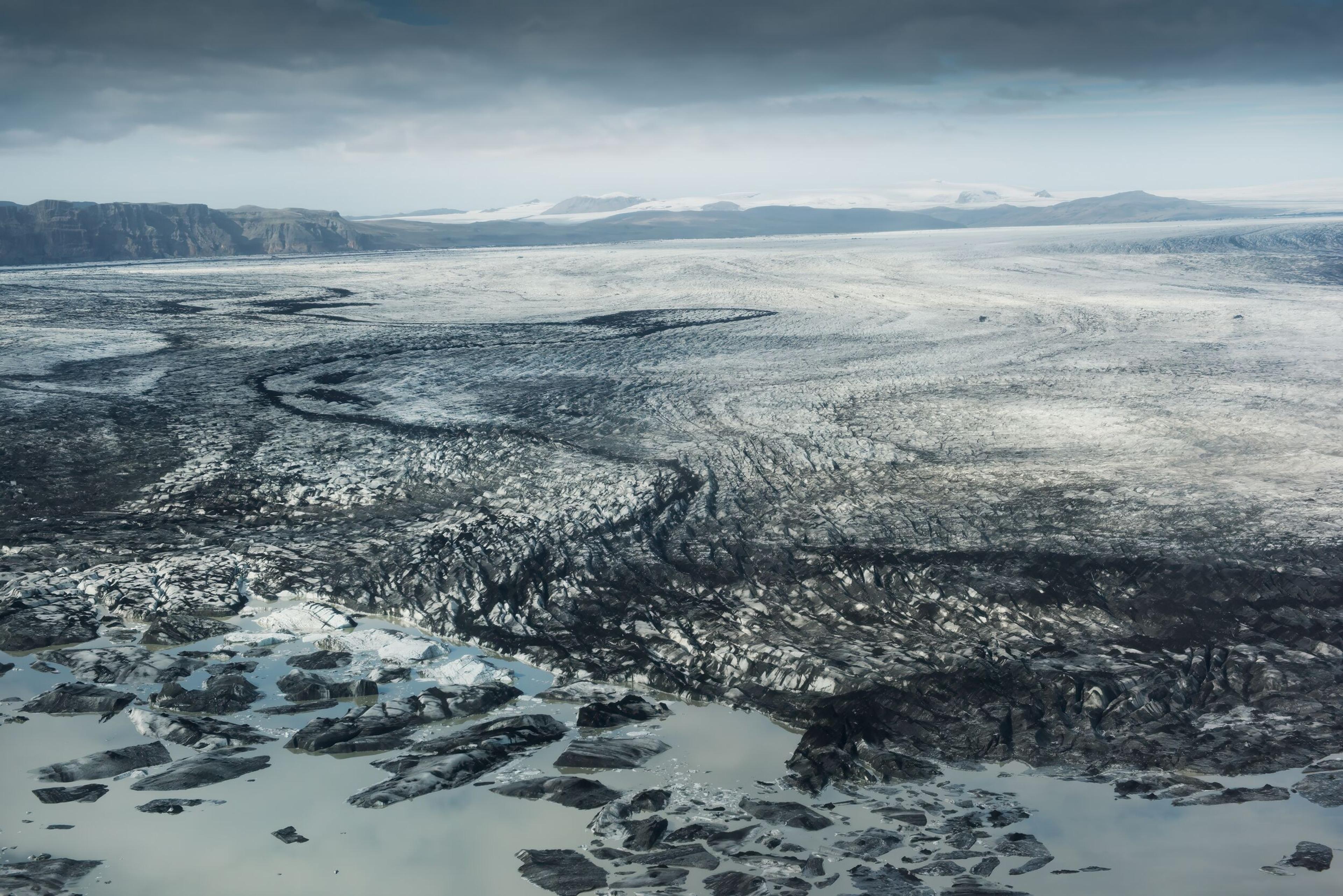 Close-up aerial view of a glacier with deep crevasses and a pattern of dark sediment lines, highlighting the dynamic and rugged texture of the ice.