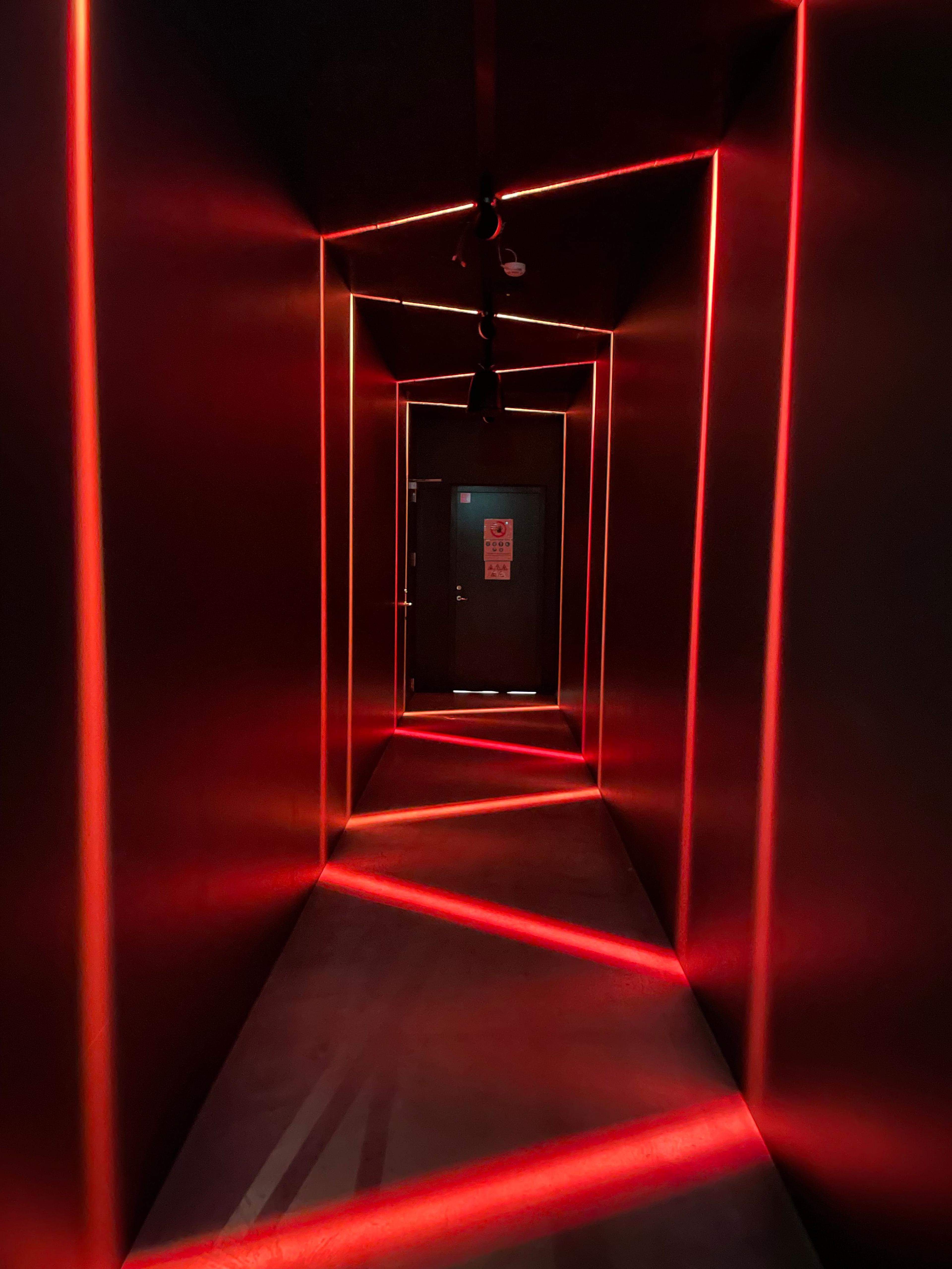 A dark hallway with red strip lights outlining its geometric shape and diagonal beams on the floor, leading to a door.
