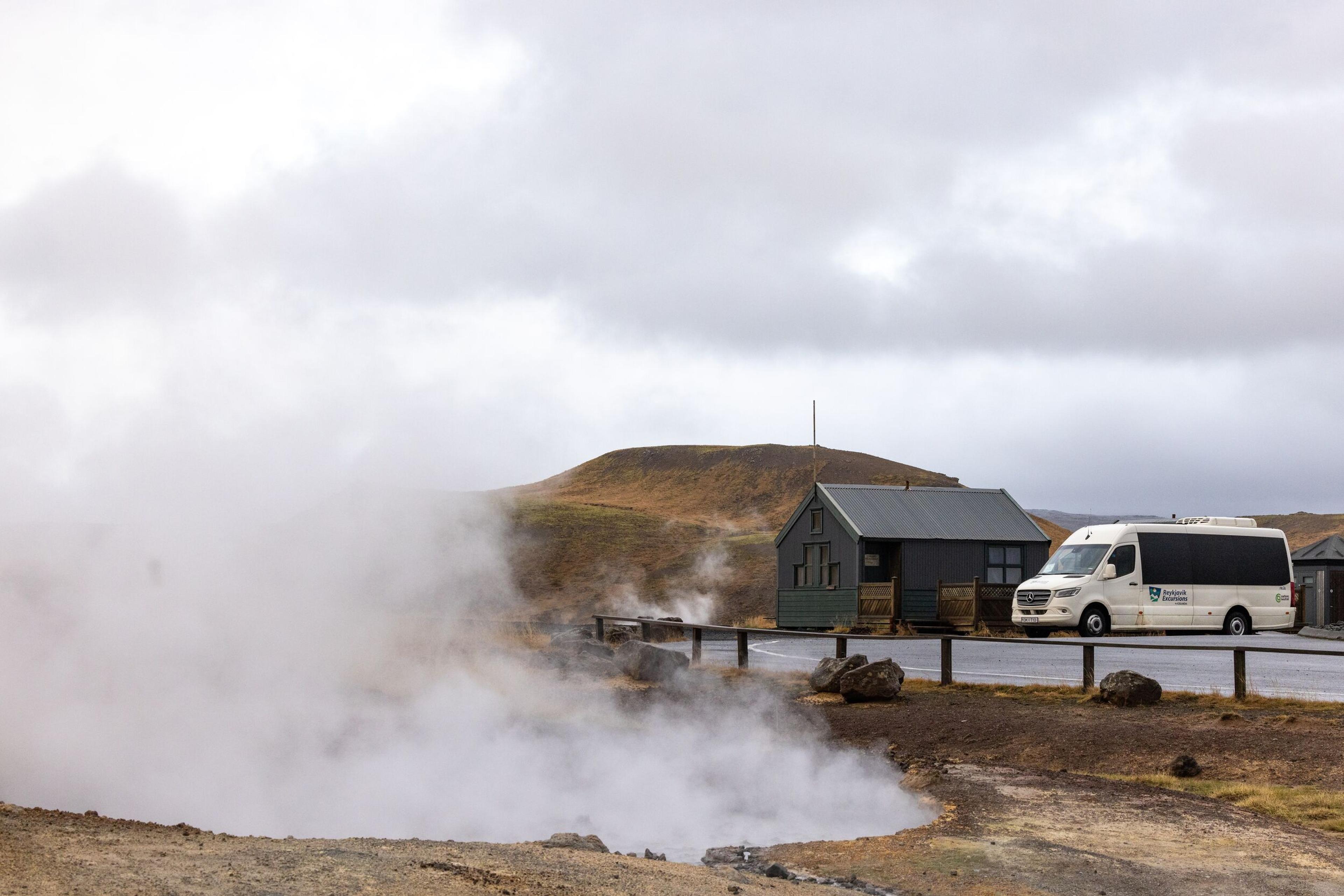 Steam rises from geothermal vents at Seltún, with a small building and a parked tour van nearby under a cloudy sky.