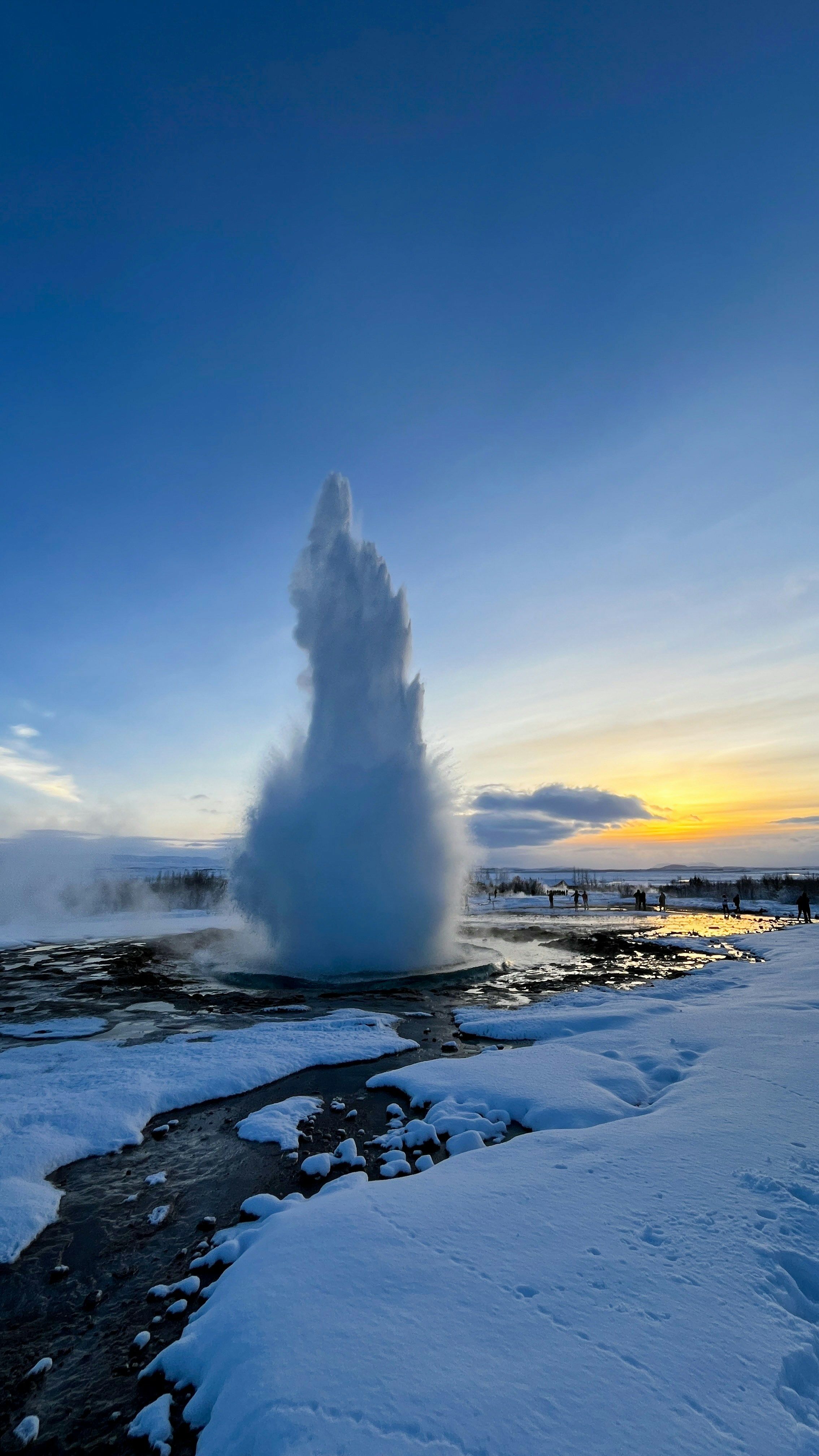 A powerful geyser eruption in Iceland shoots water high into the winter sky, set against a serene sunset and surrounded by a snowy landscape.
