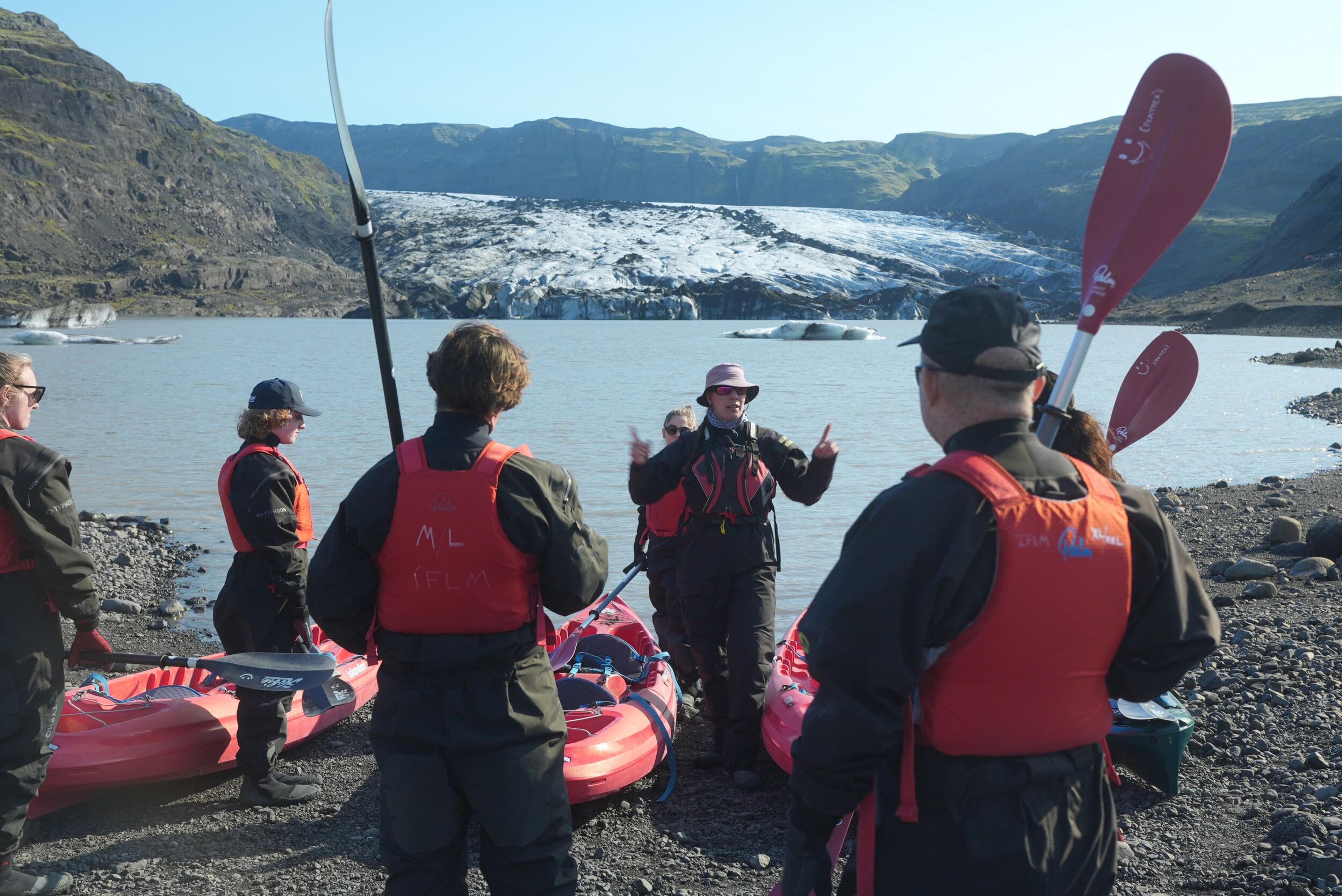 A guide briefs kayakers in dry suits and life vests on a glacial lagoon shore, with a large glacier behind them.