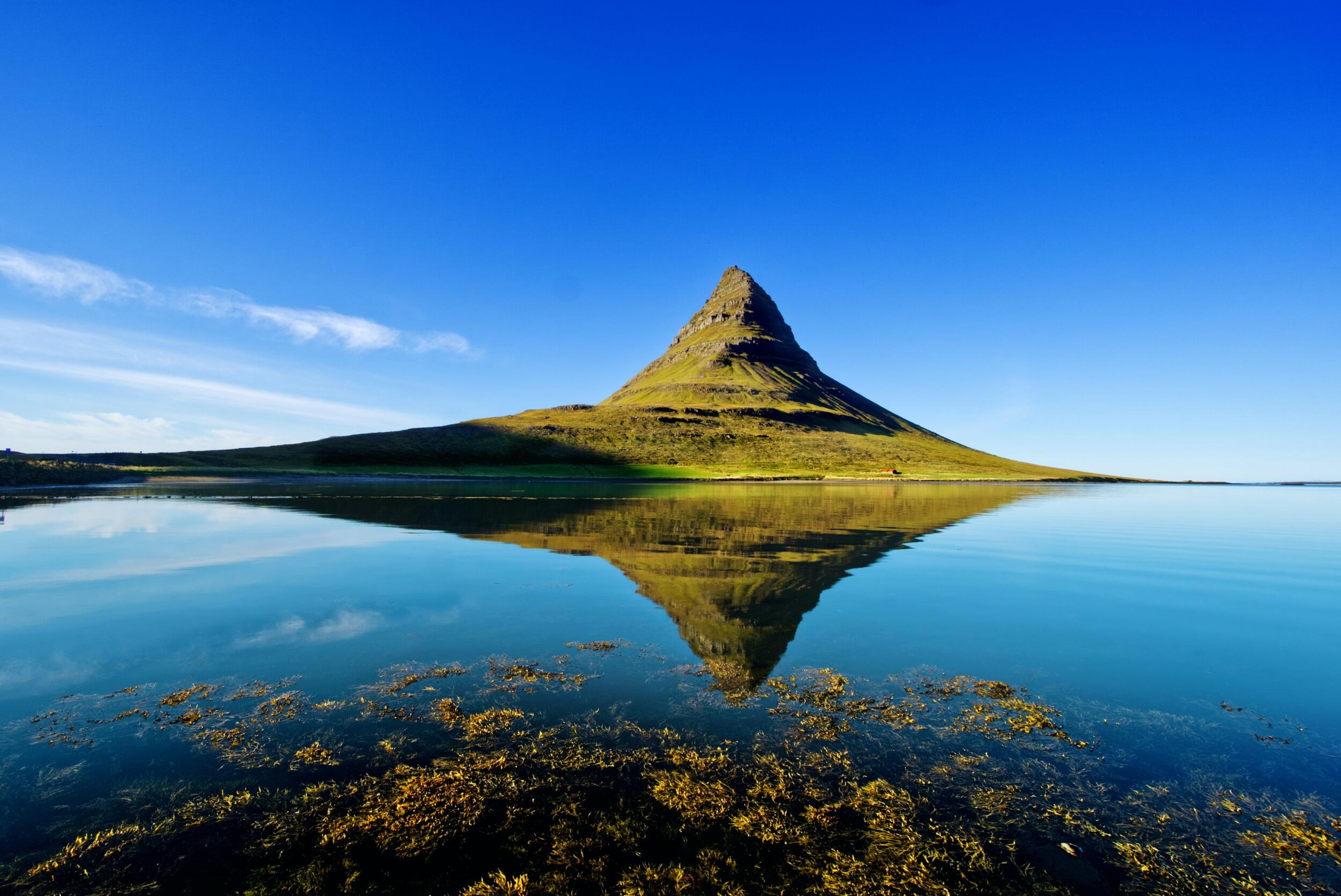 Kirkjufell mountain in Iceland reflects perfectly in the still waters below, with a clear blue sky framing its iconic peak.