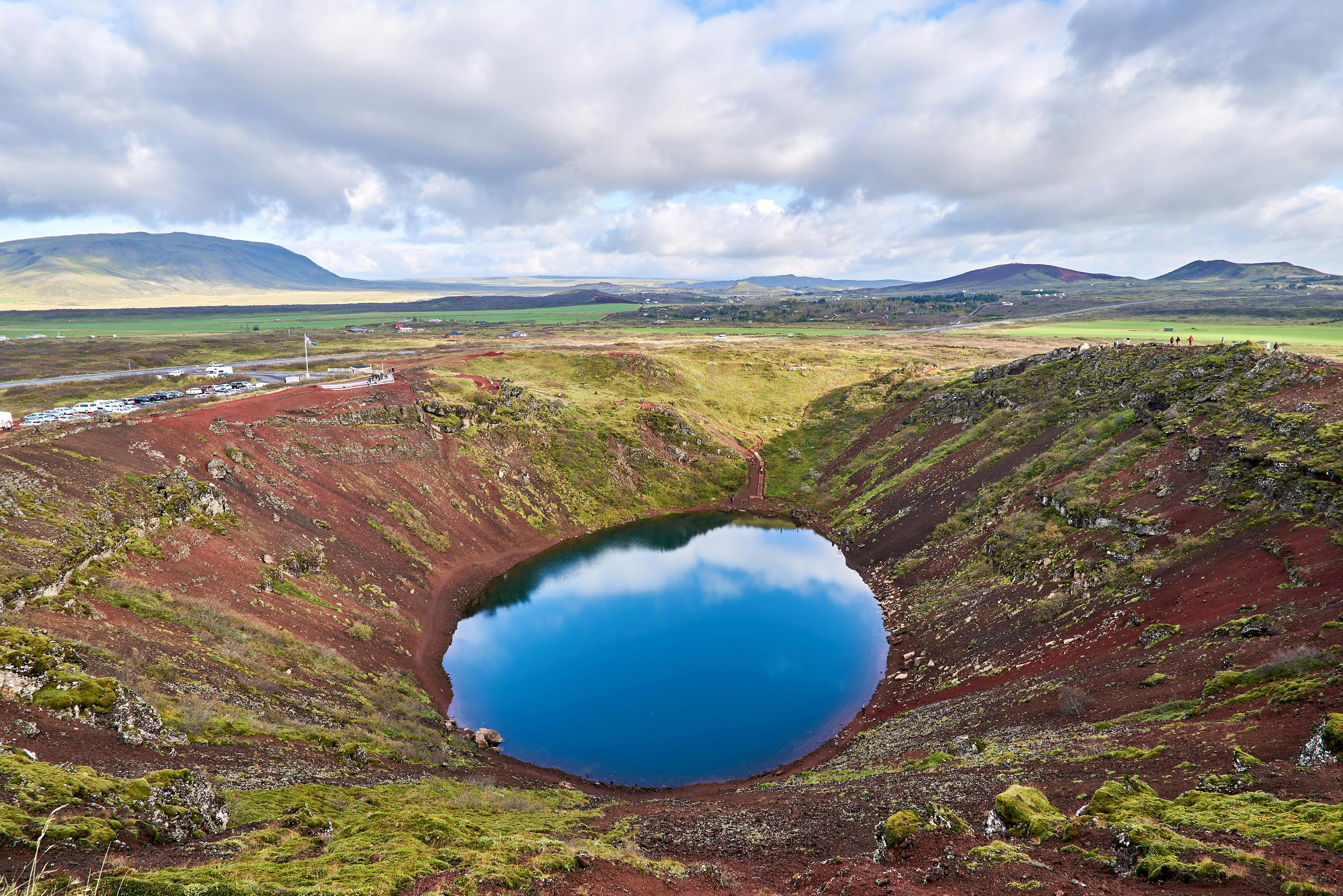 A vibrant blue volcanic crater lake nestled within a rich, red-earthed depression, with a panoramic view of the expansive, green Icelandic landscape under a cloud-strewn sky.