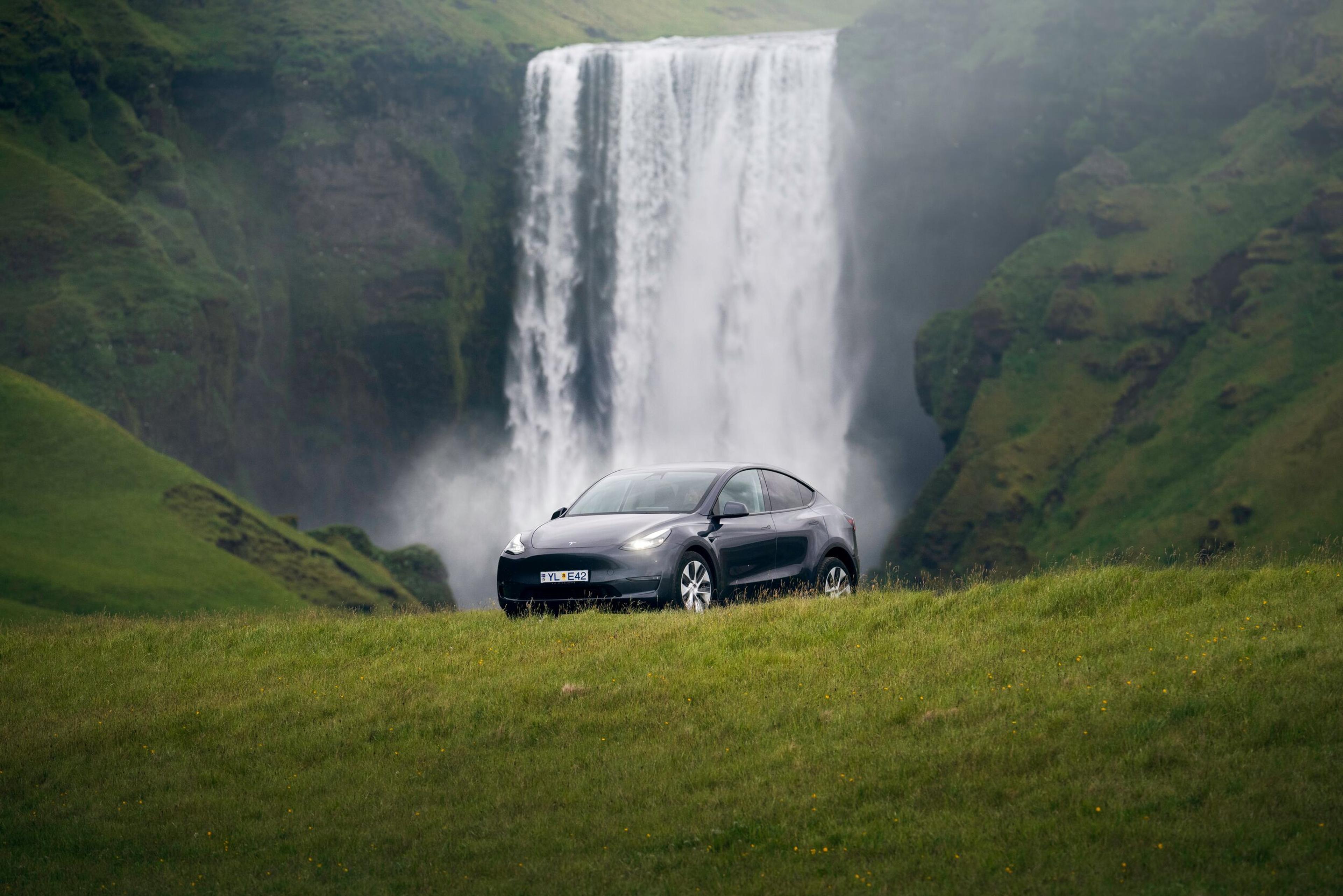 A black Tesla sits parked in front of a majestic waterfall in Iceland, capturing the beauty of an Iceland car rental adventure.