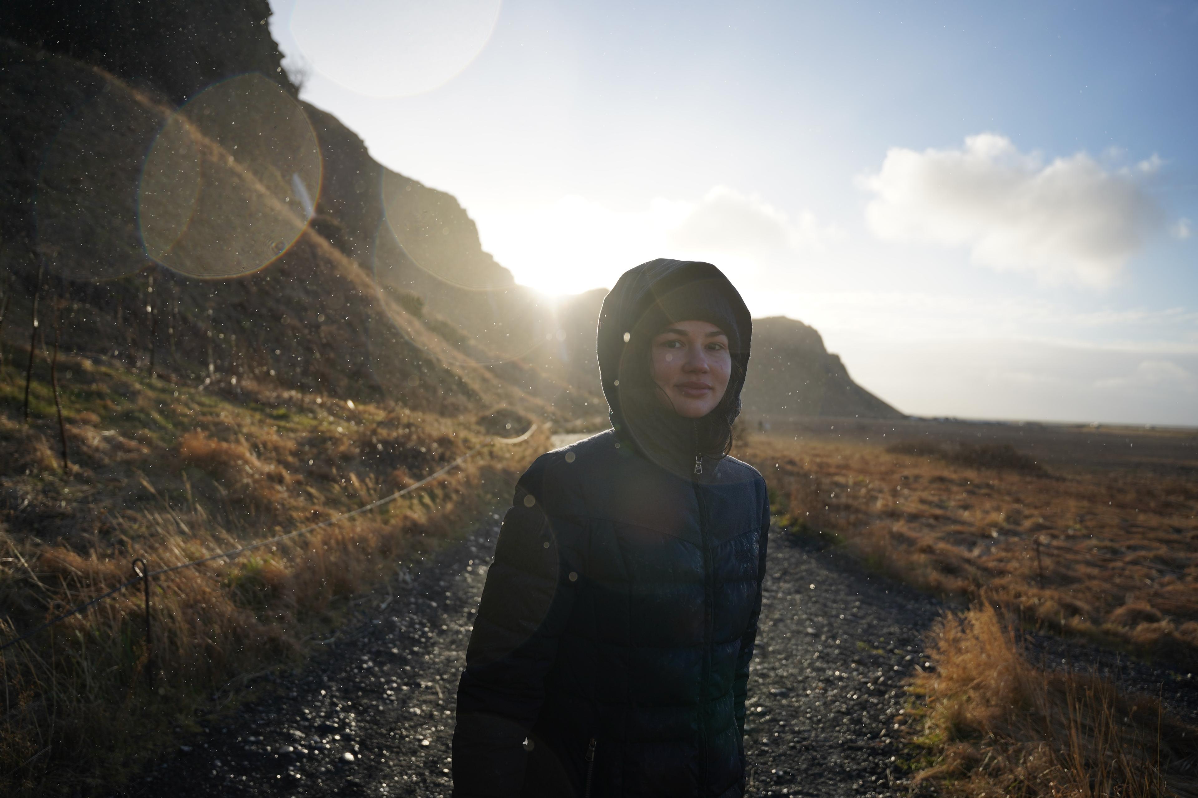 A woman with a hood over her hat, smiling at the camera, caught in a light shower yet illuminated by sunshine.
