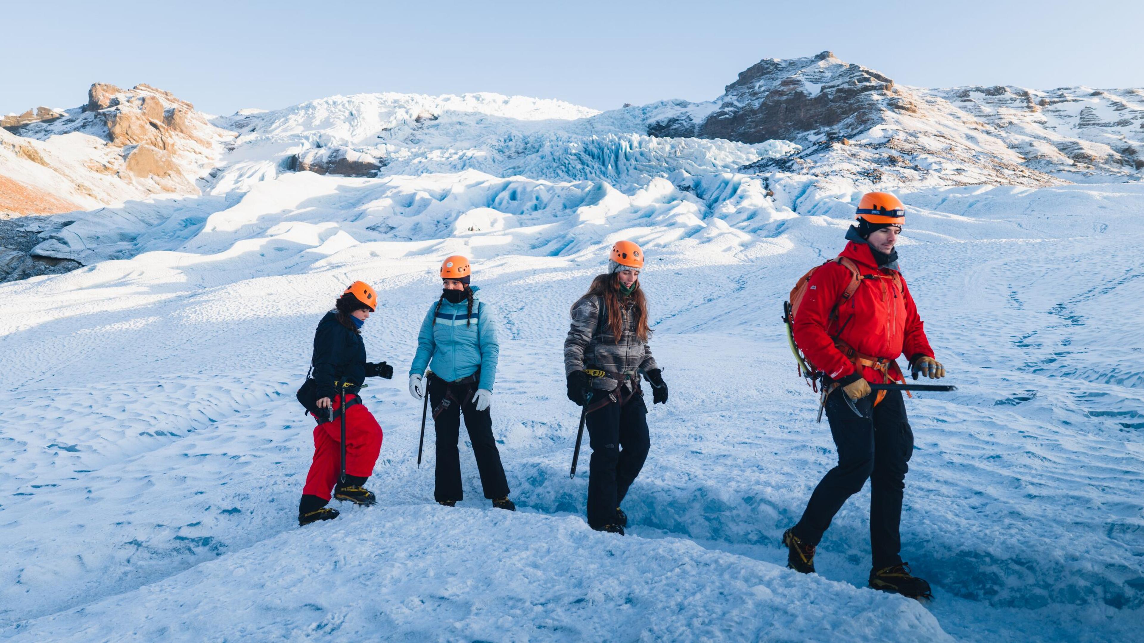 Four people in helmets and winter gear glacier hiking on an icy landscape.