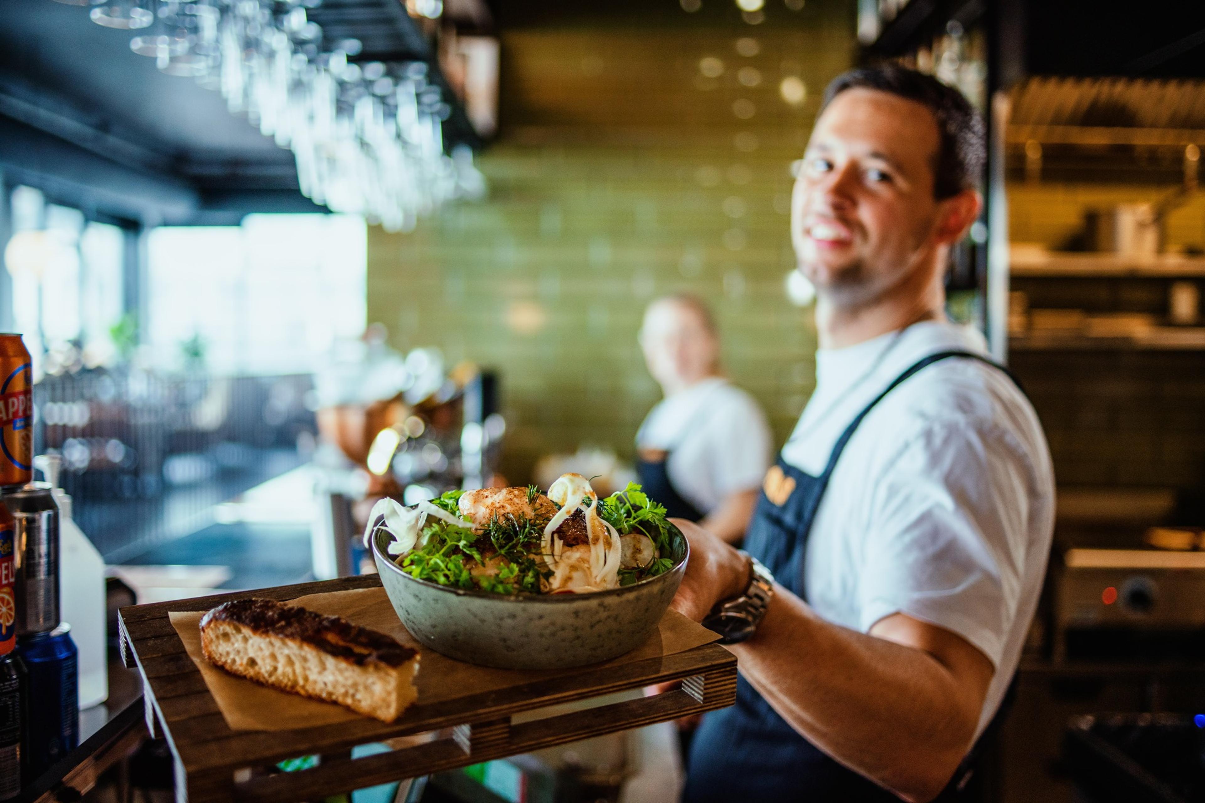 Server presenting a fresh, vibrant salad with grilled bread at Borg29 Food Hall, smiling in a welcoming, modern dining environment.