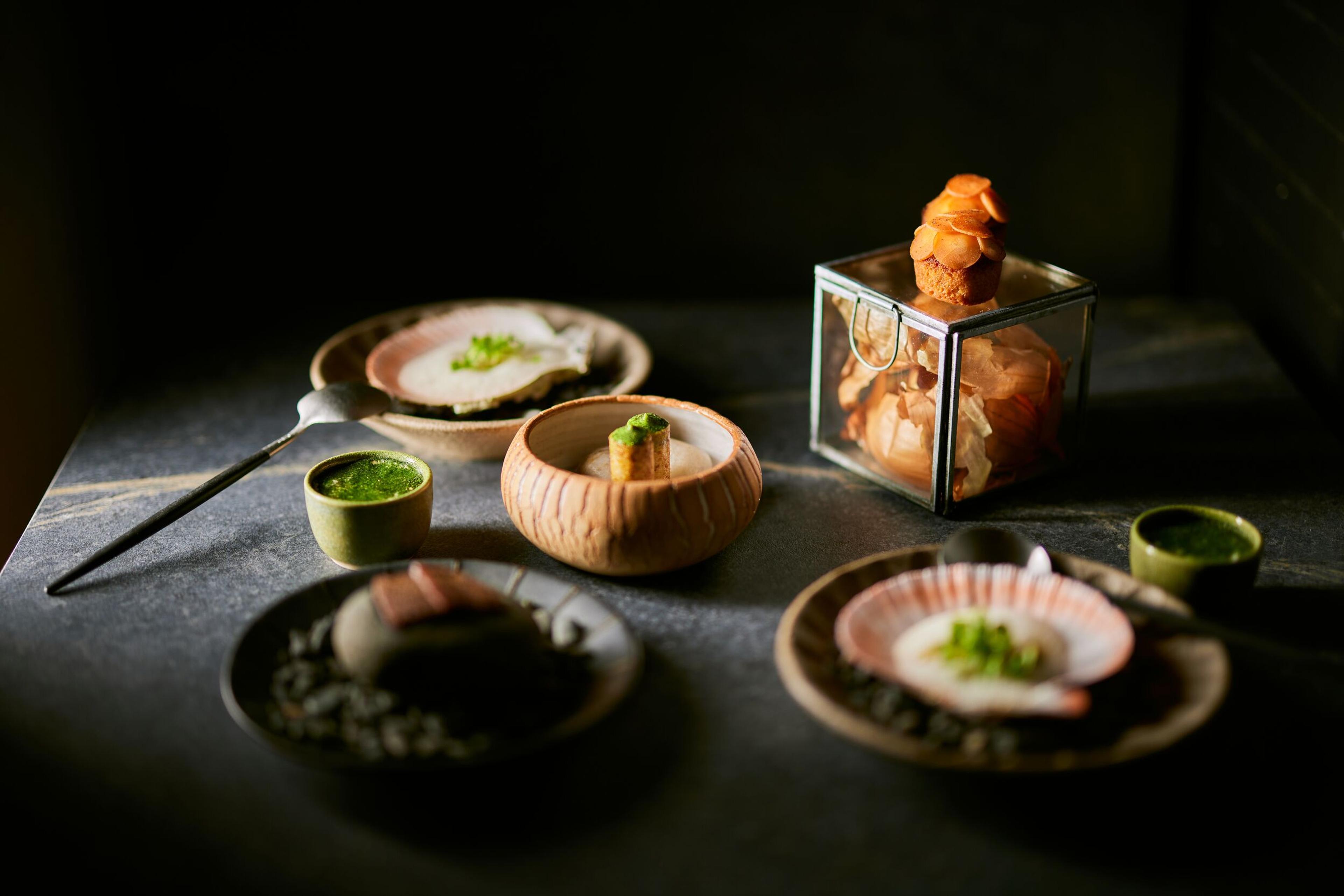 Multiple exquisitely plated small dishes, including a scallop and a glass box with treats, on a dark table.