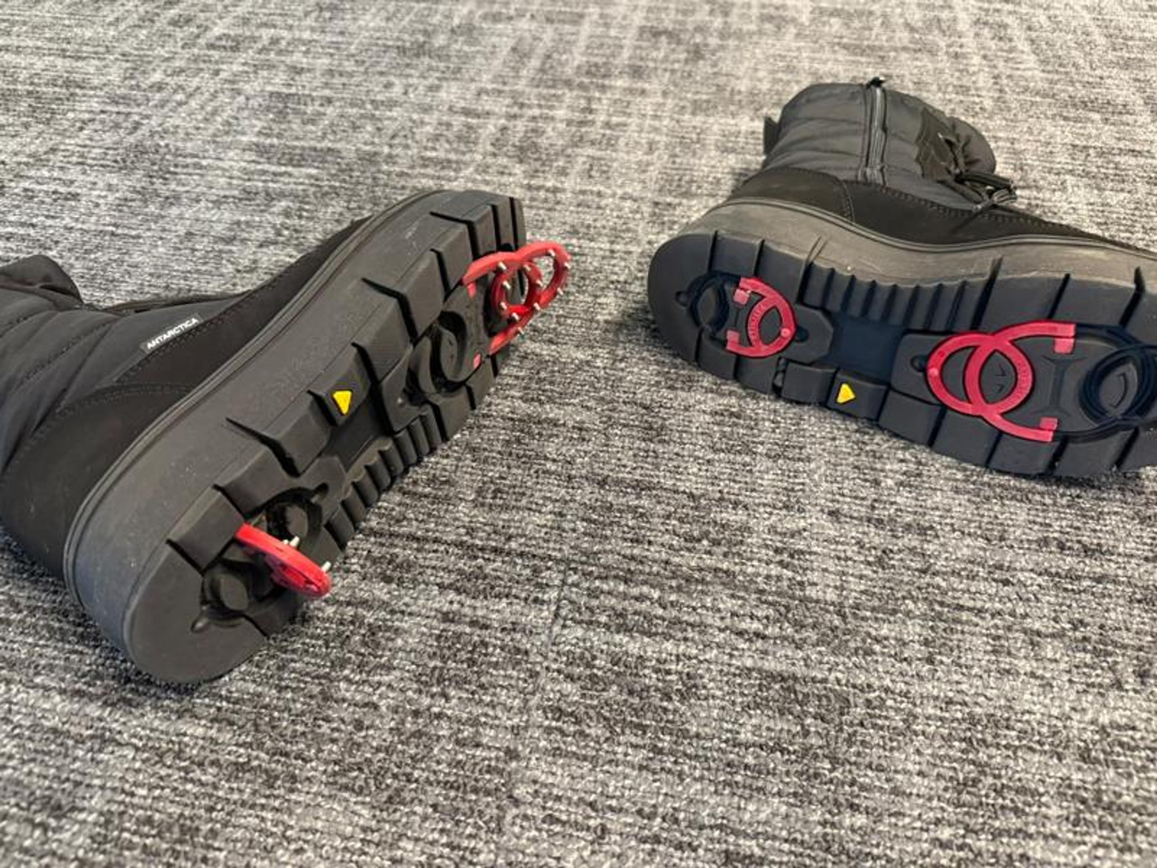 Two black winter boots on a grey carpet; one boot shows deployed red ice grips on its sole, while the other displays recessed red traction circles.