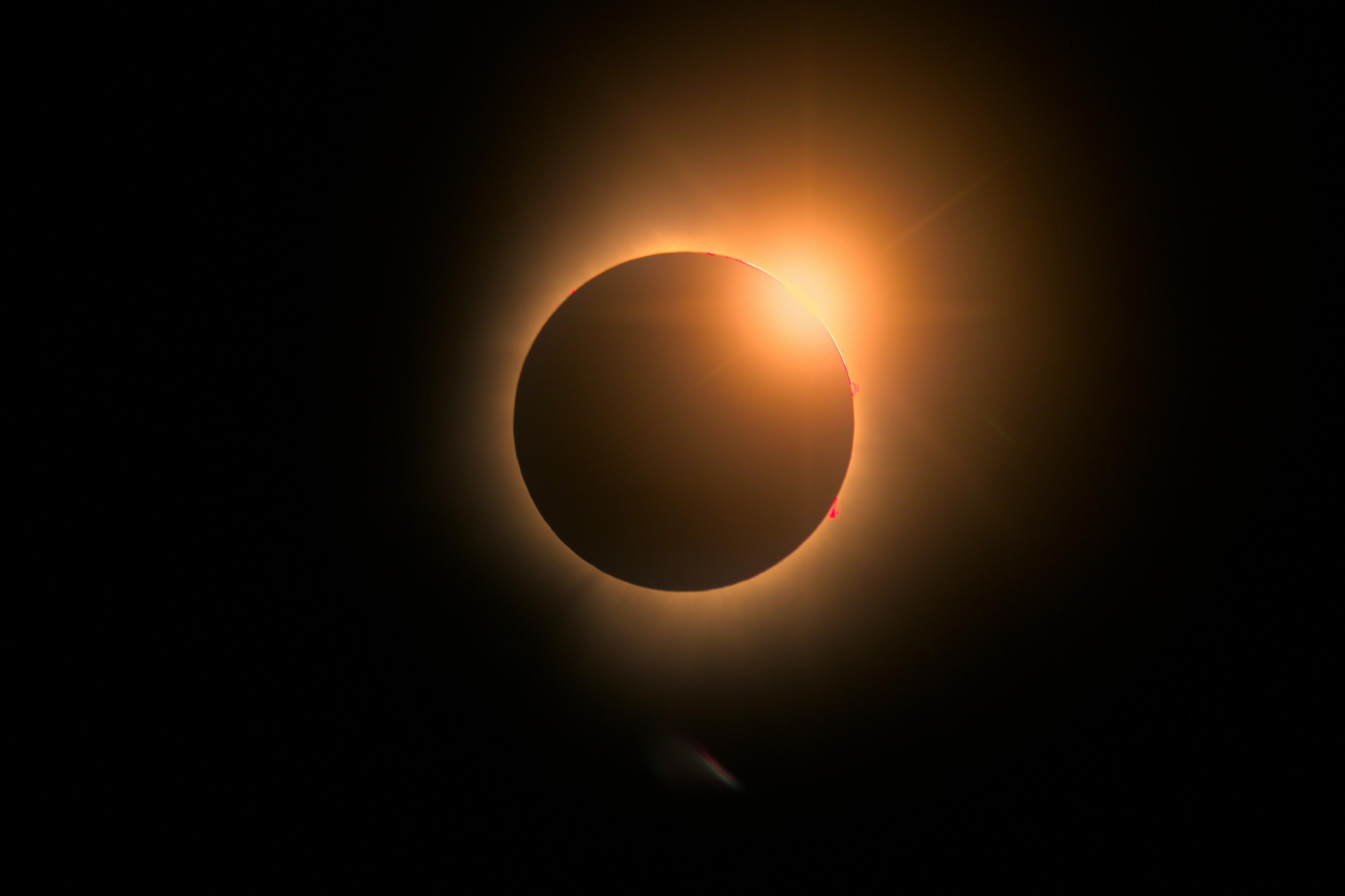 Silhouette of an airplane flying across a bright crescent sun during a solar eclipse, set against an orange sky.