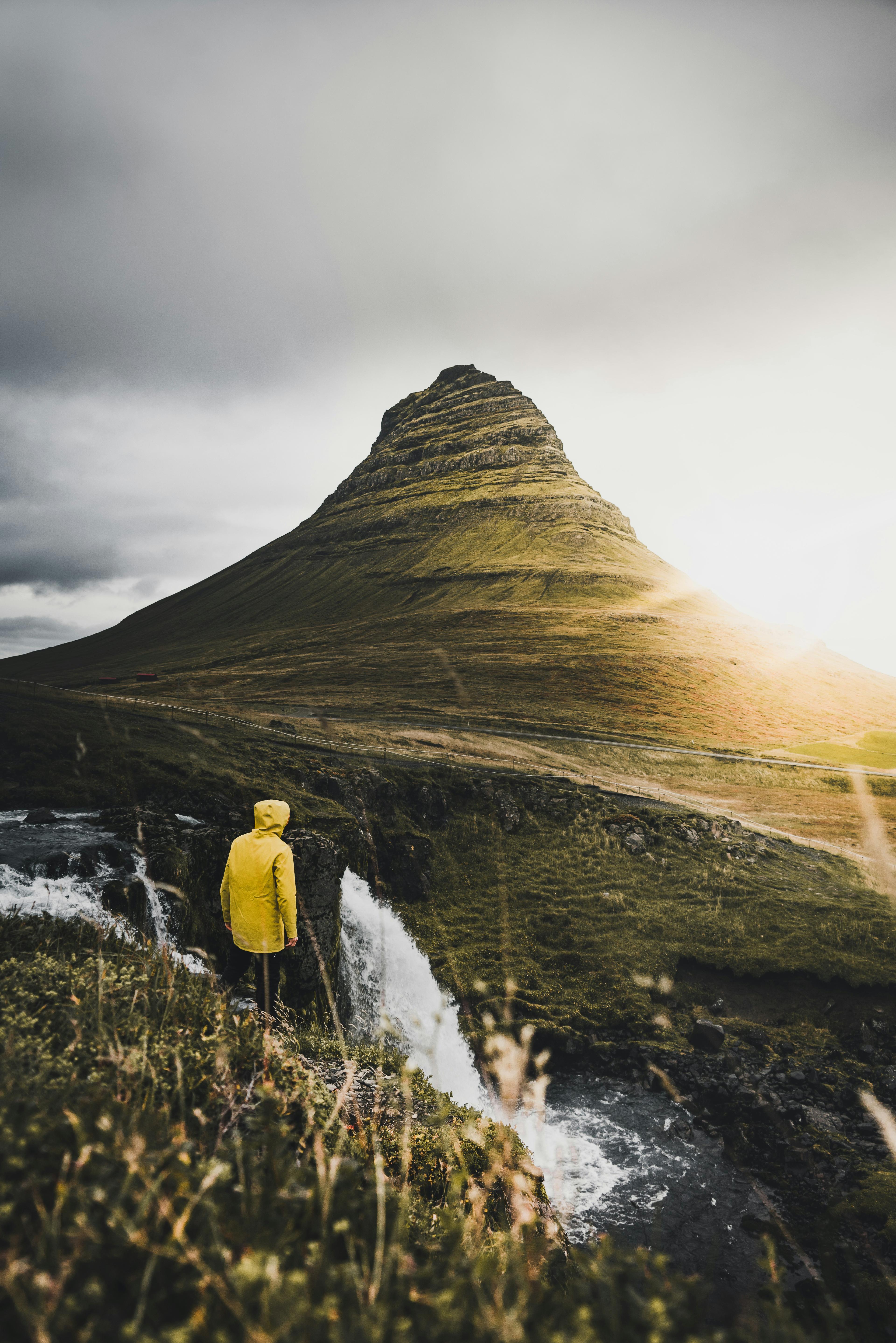 A person in a bright yellow jacket stands on a hill overlooking a waterfall, facing a dramatic, sharply pointed mountain bathed in the warm glow of a setting sun.