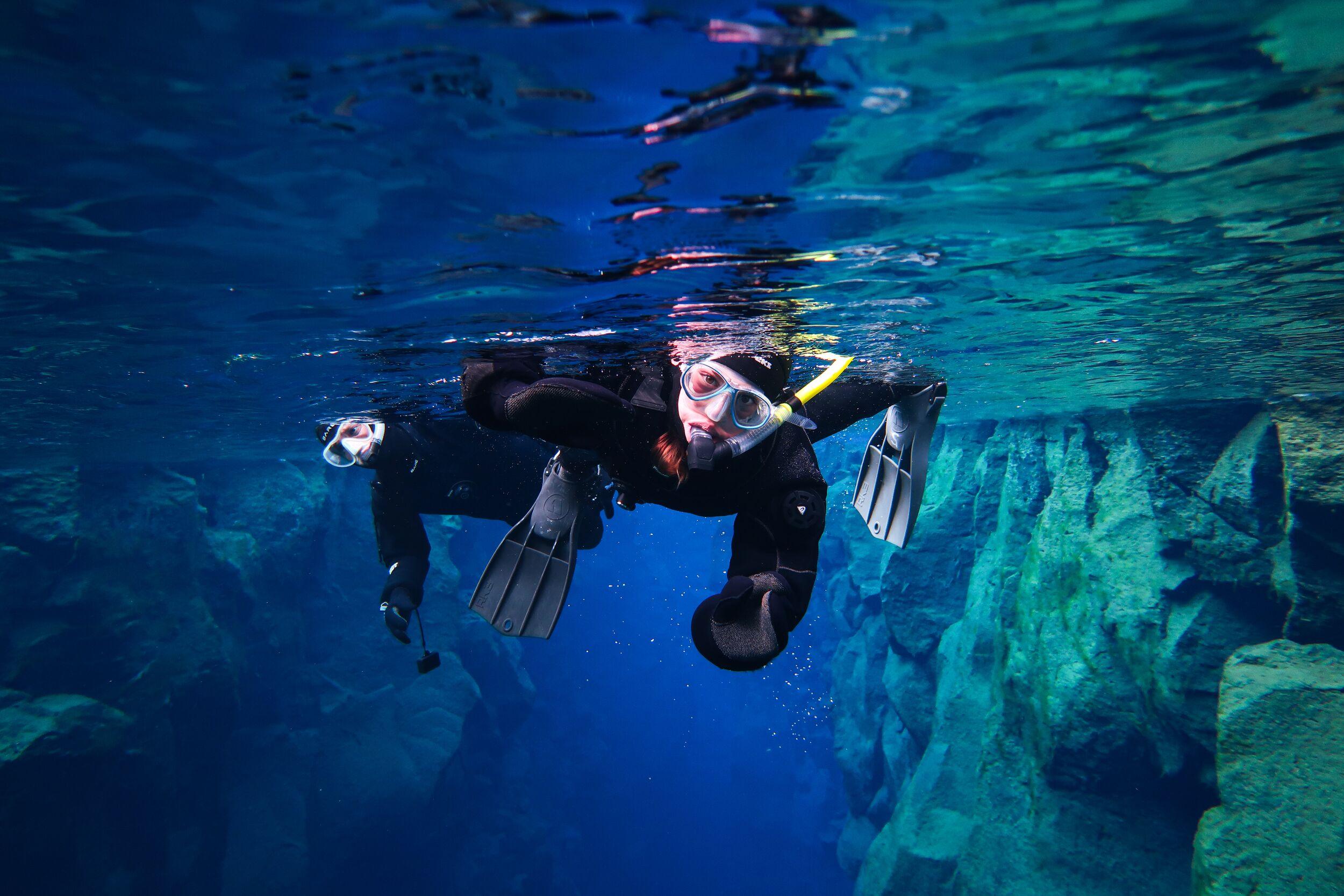Two people in wetsuits, masks, and fins swim through a clear blue underwater canyon.