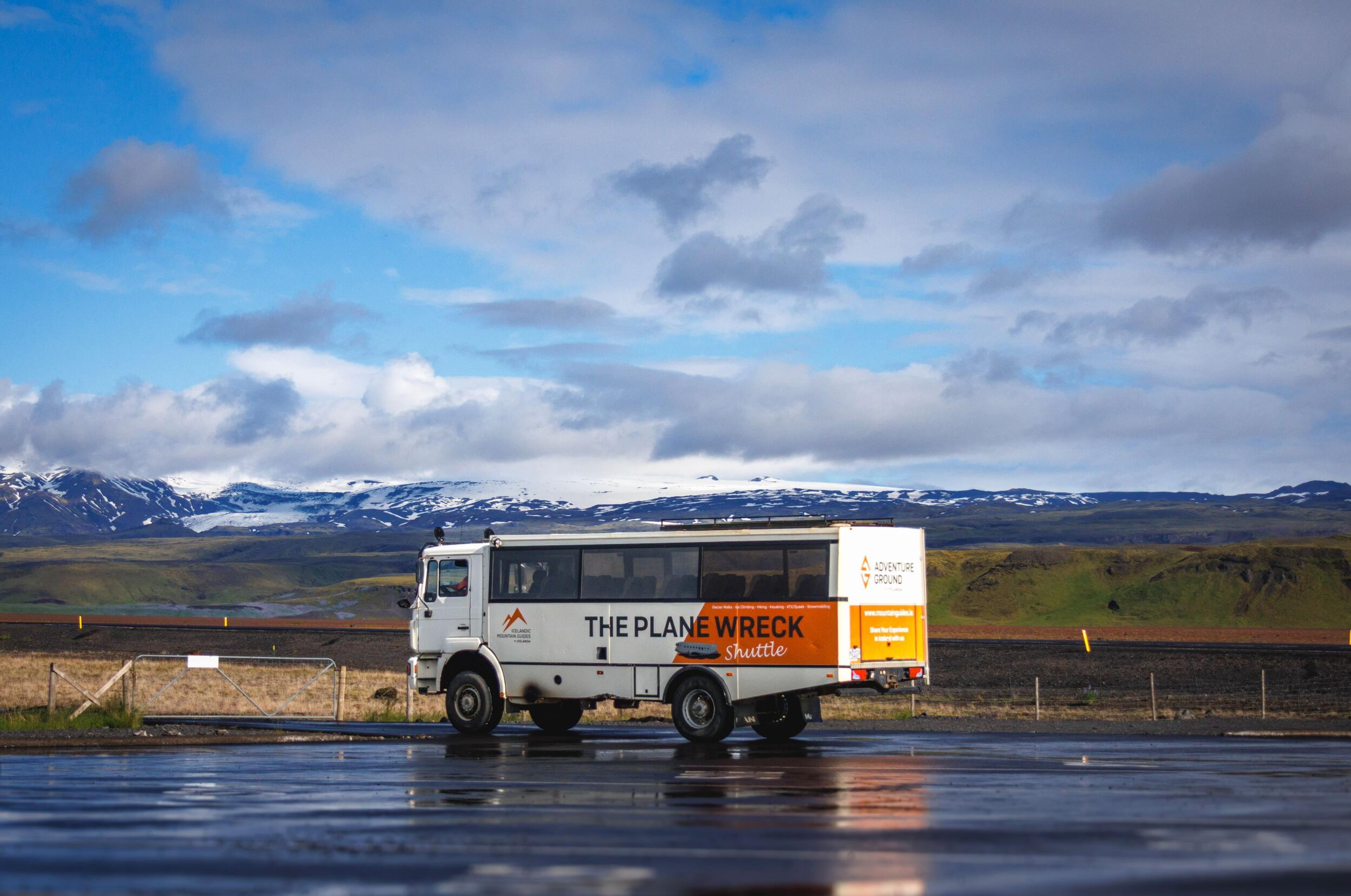 A white shuttle bus for "The Plane Wreck" tour on a wet road with snow-capped mountains in the background.