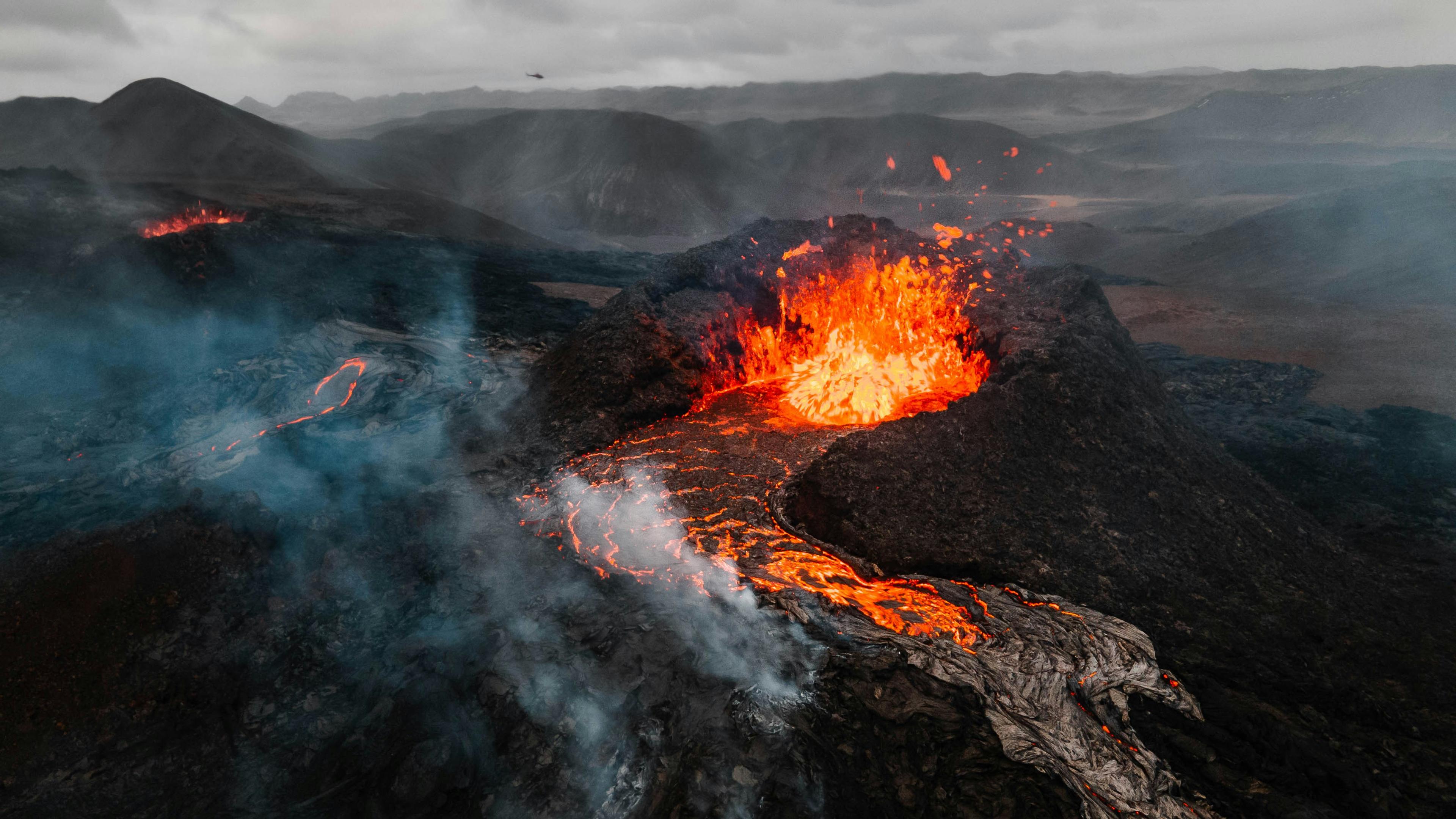 Aerial shot of a fiery volcanic eruption with molten lava flows and smoke in a mountainous terrain.