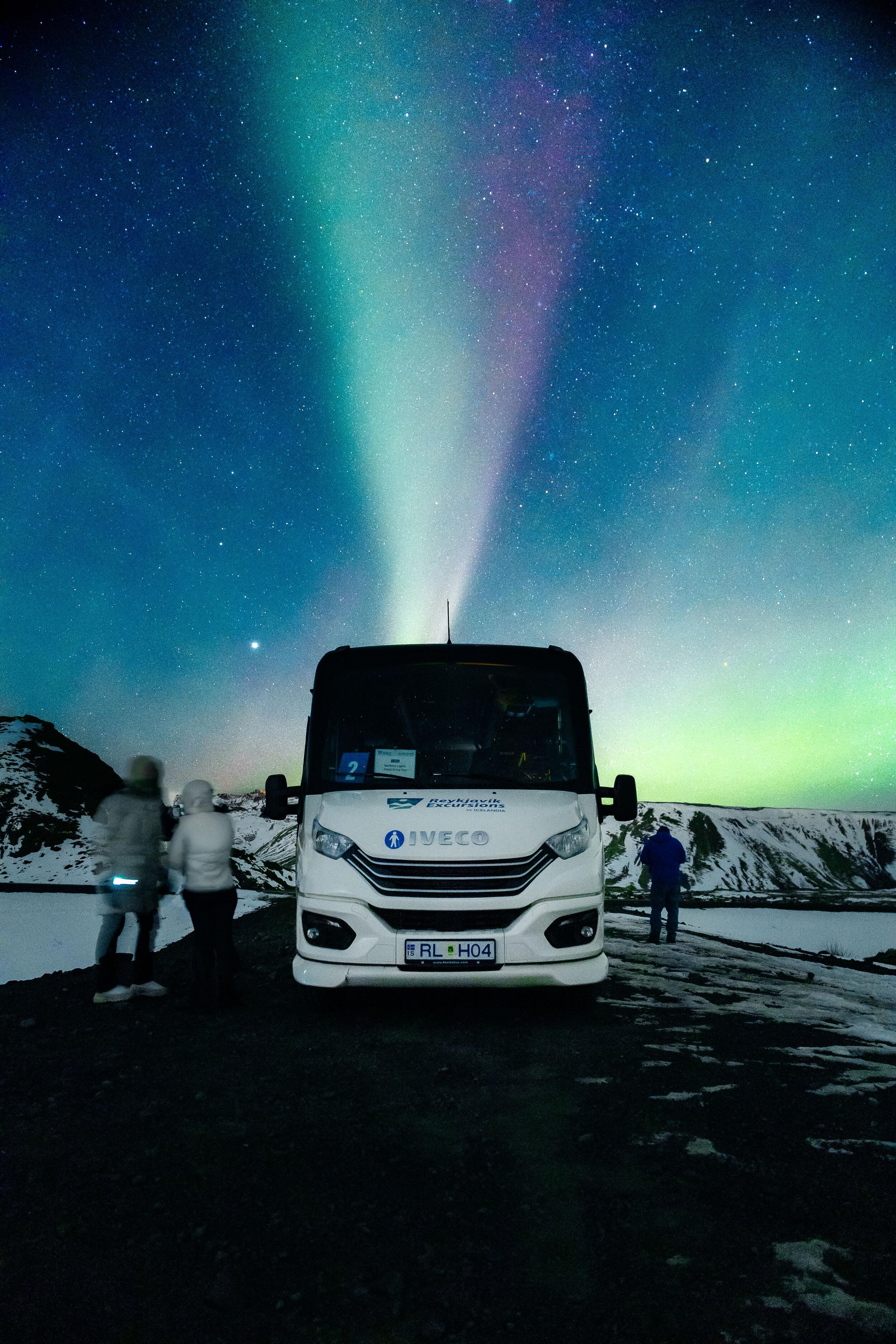 A white tour bus and people under a vibrant green and purple aurora in a starry, snowy landscape.