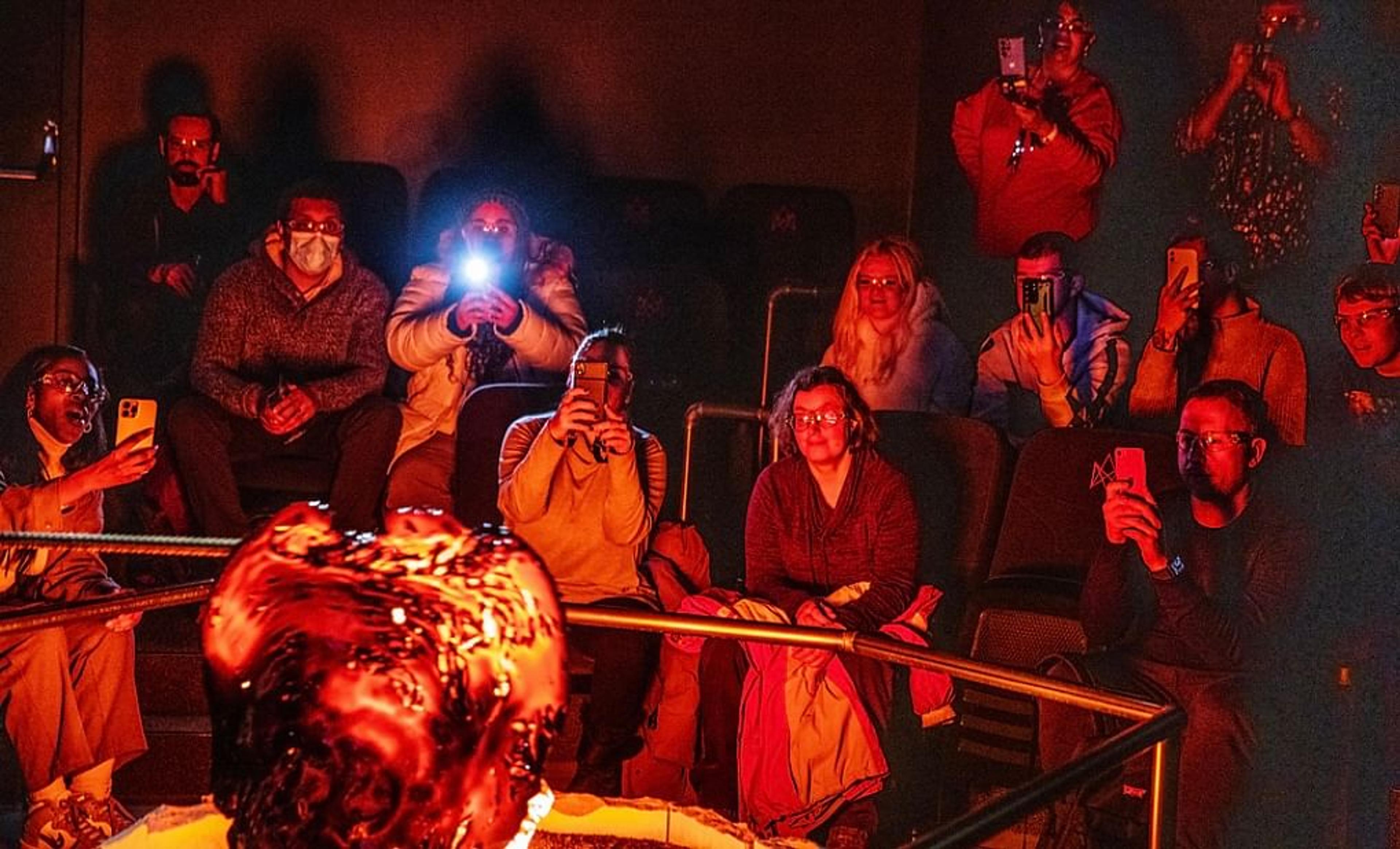 A group of people in a dark, red-lit room capturing photos of a glowing, molten object with their phones.