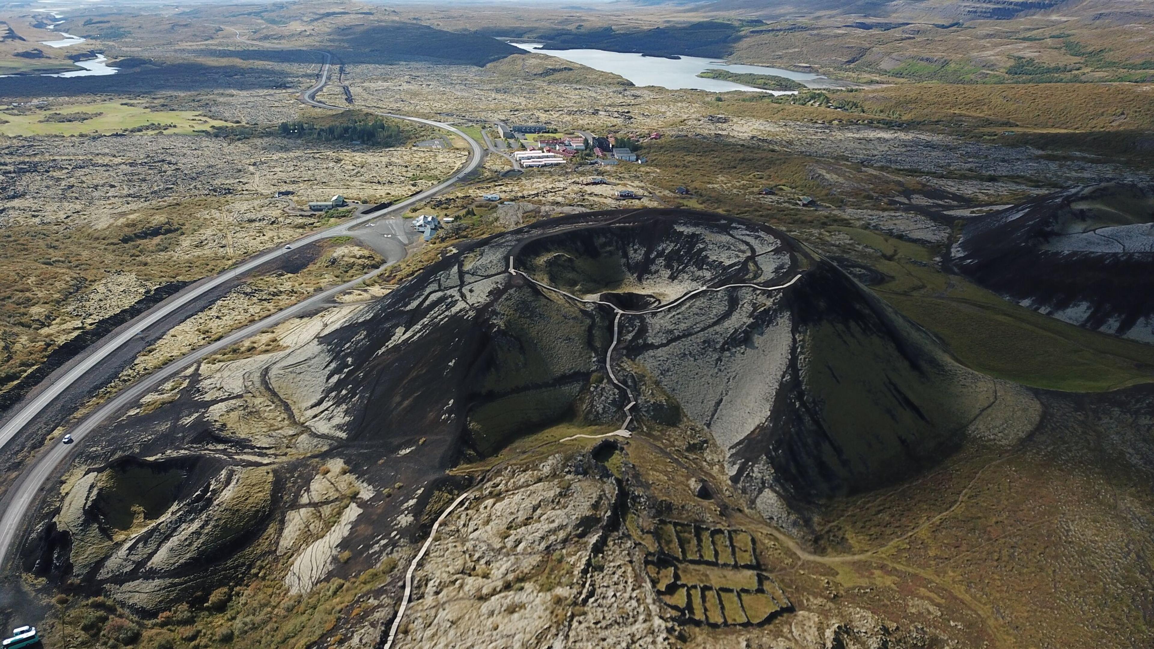 Aerial view of Grábrók crater, showcasing its volcanic cone with walking paths leading to the summit, surrounded by barren lava fields, grassy patches, and distant lakes in Iceland's rugged landscape.