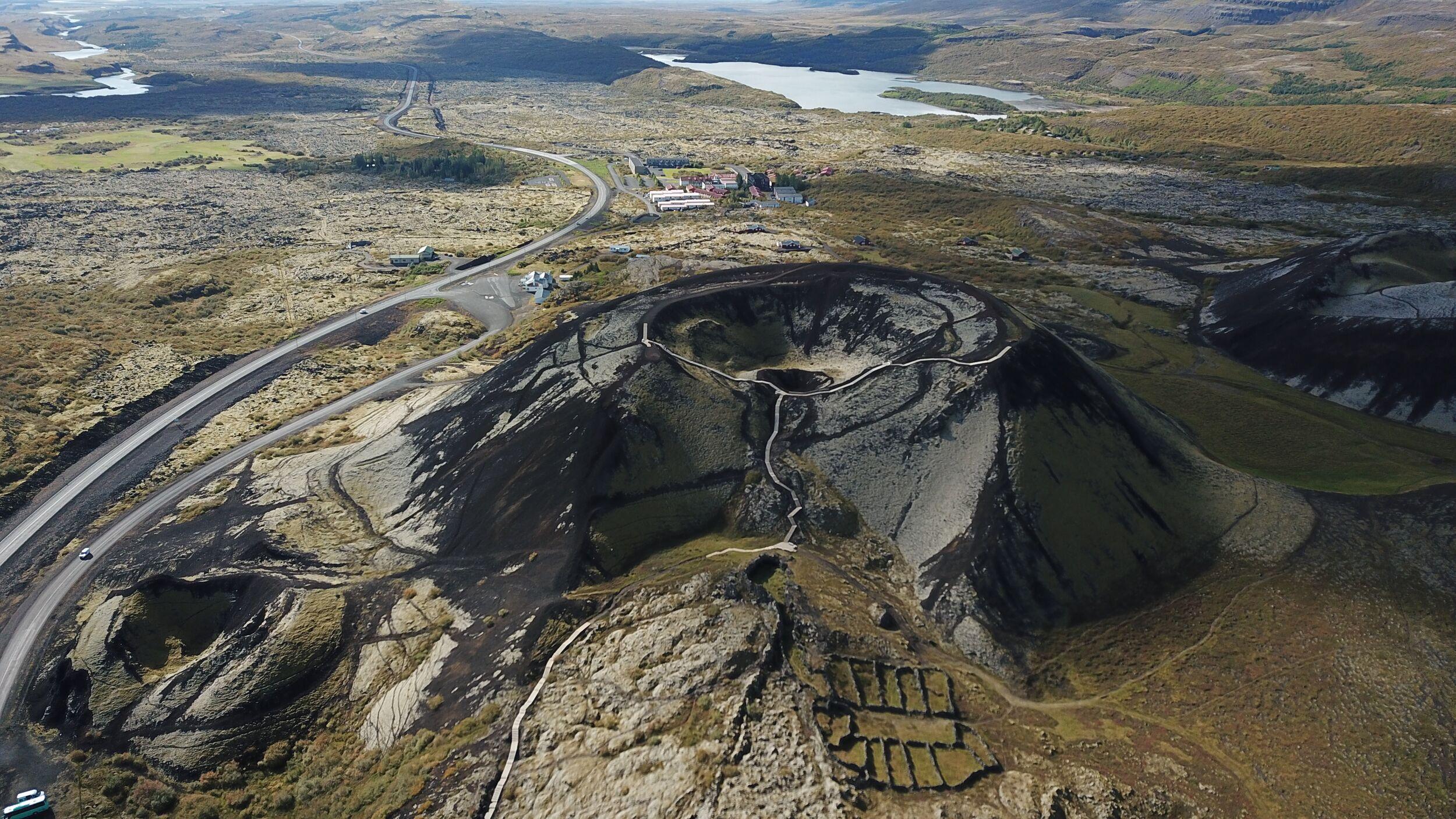 Aerial view of Grábrók crater, showcasing its volcanic cone with walking paths leading to the summit, surrounded by barren lava fields, grassy patches, and distant lakes in Iceland's rugged landscape.