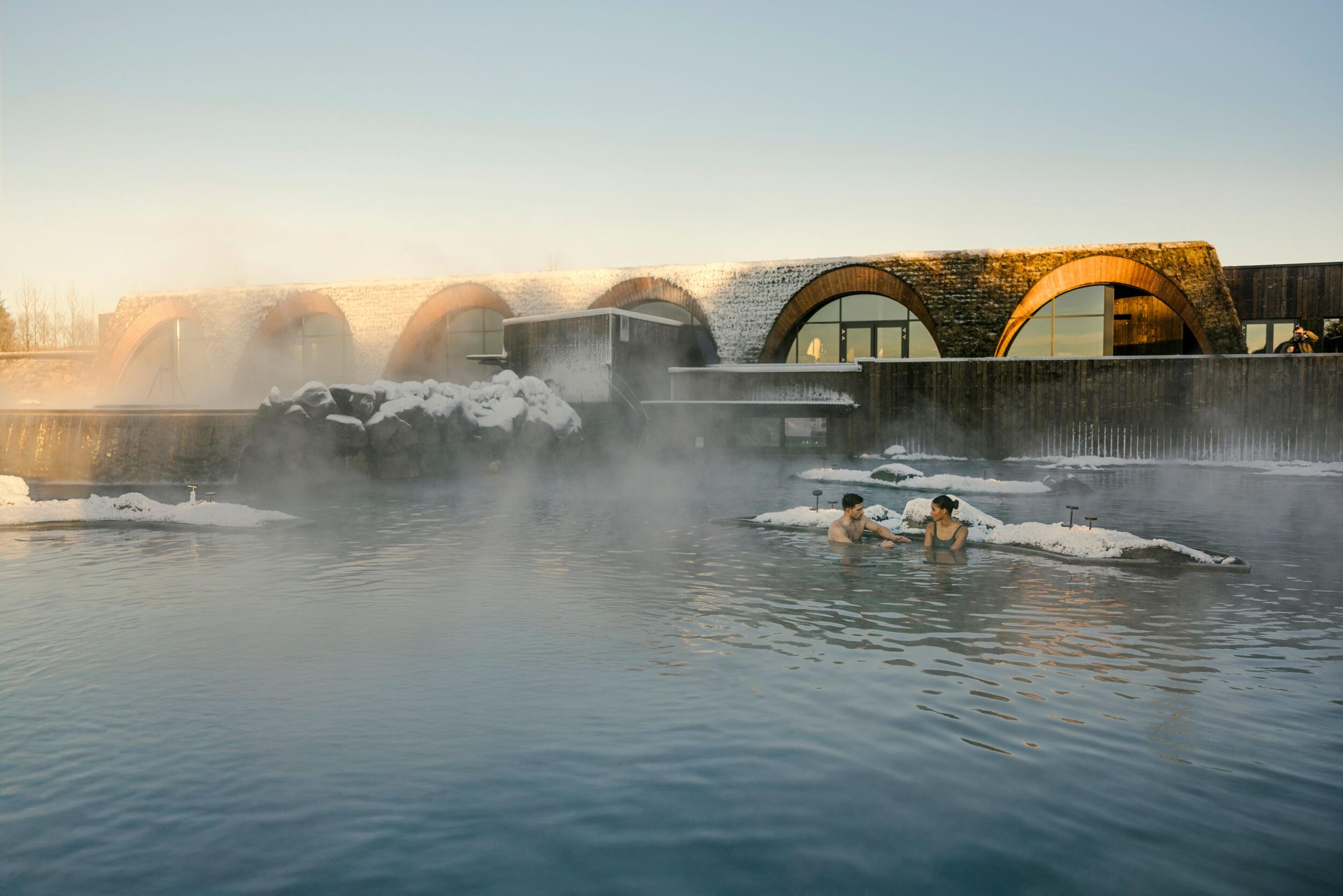 Two people relax in a steaming blue geothermal pool with a modern, snow-covered building in the background.