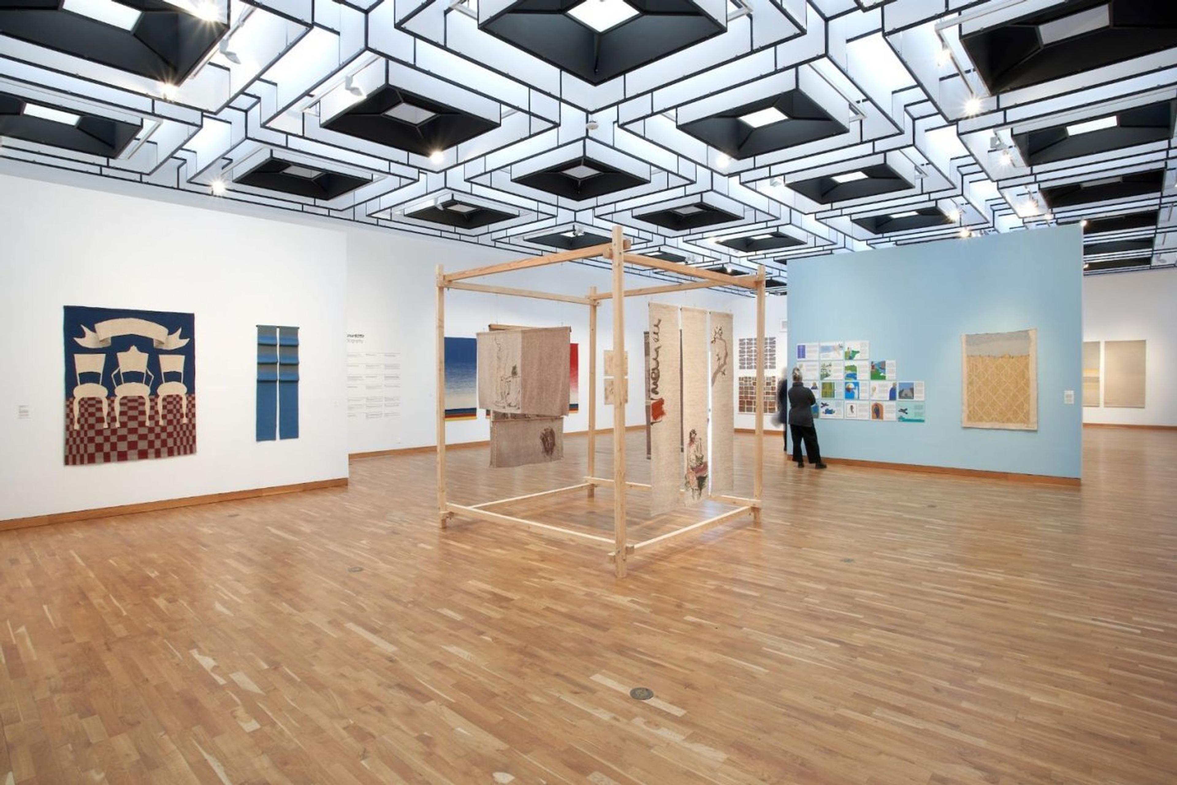 Modern art gallery with textile and mixed-media displays on wooden floors under geometric ceiling lights.