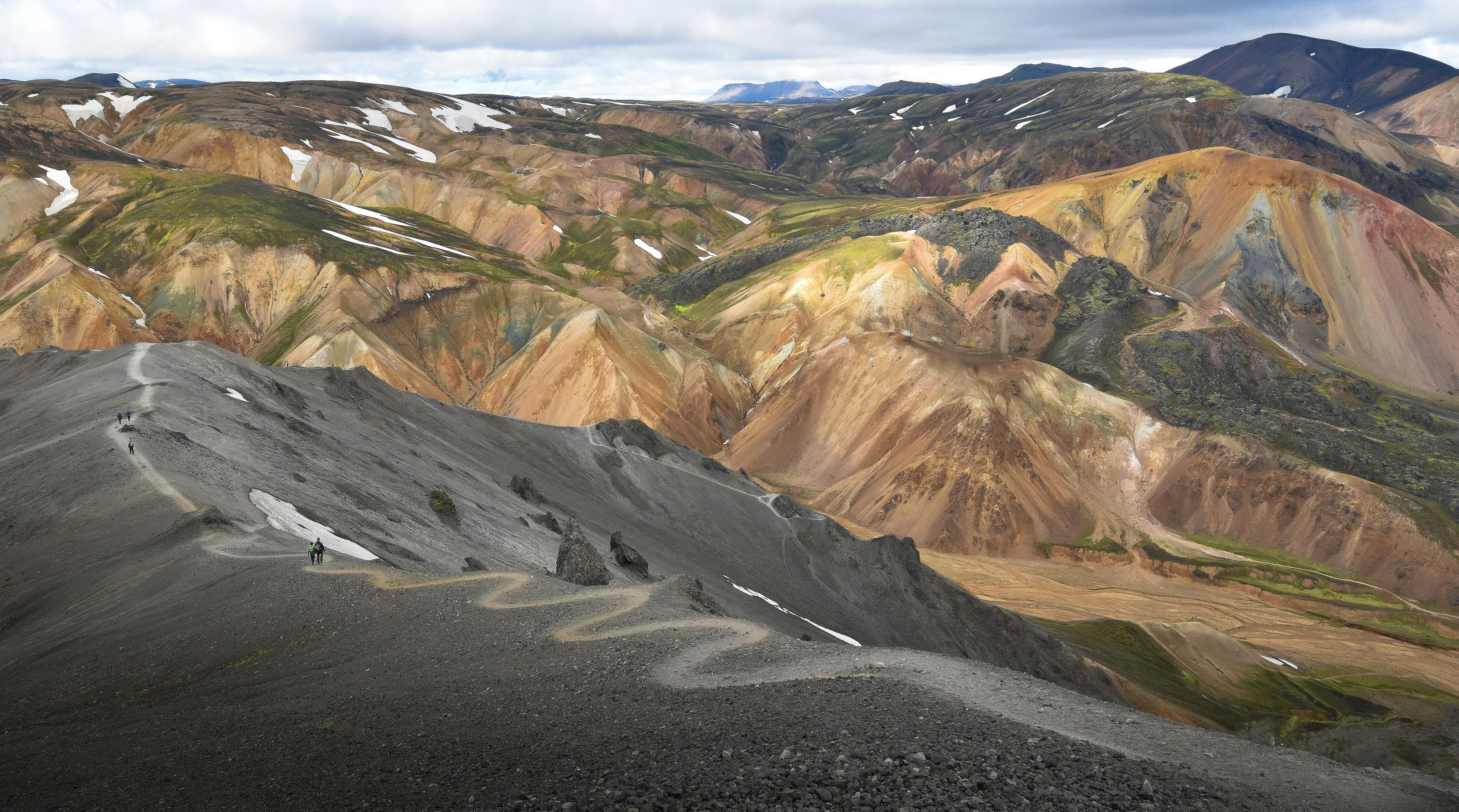  Hikers follow a steep, zigzagging trail on a gray ridge surrounded by the vibrant, volcanic terrain of Landmannalaugar in Fjallabak.