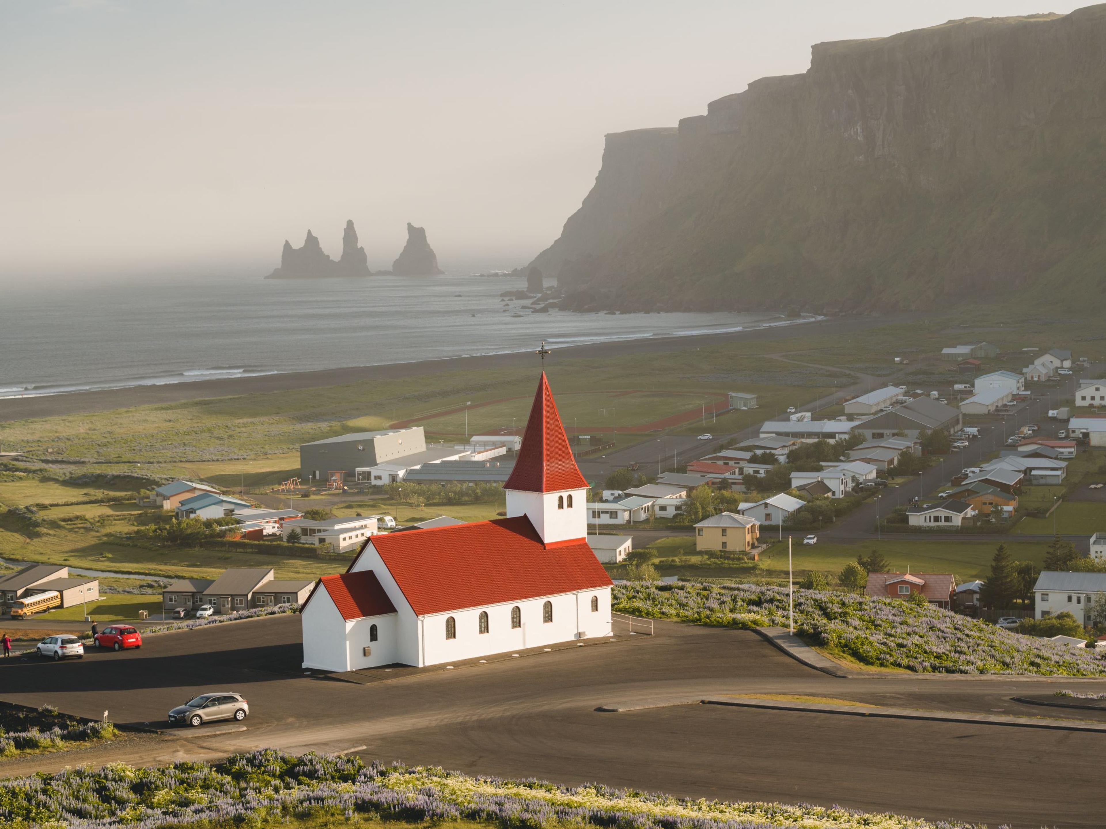 The quaint Vík village, characterized by its iconic red-roofed church standing prominently in the center, with the majestic Reynisdrangar sea stacks piercing the horizon in the backdrop.