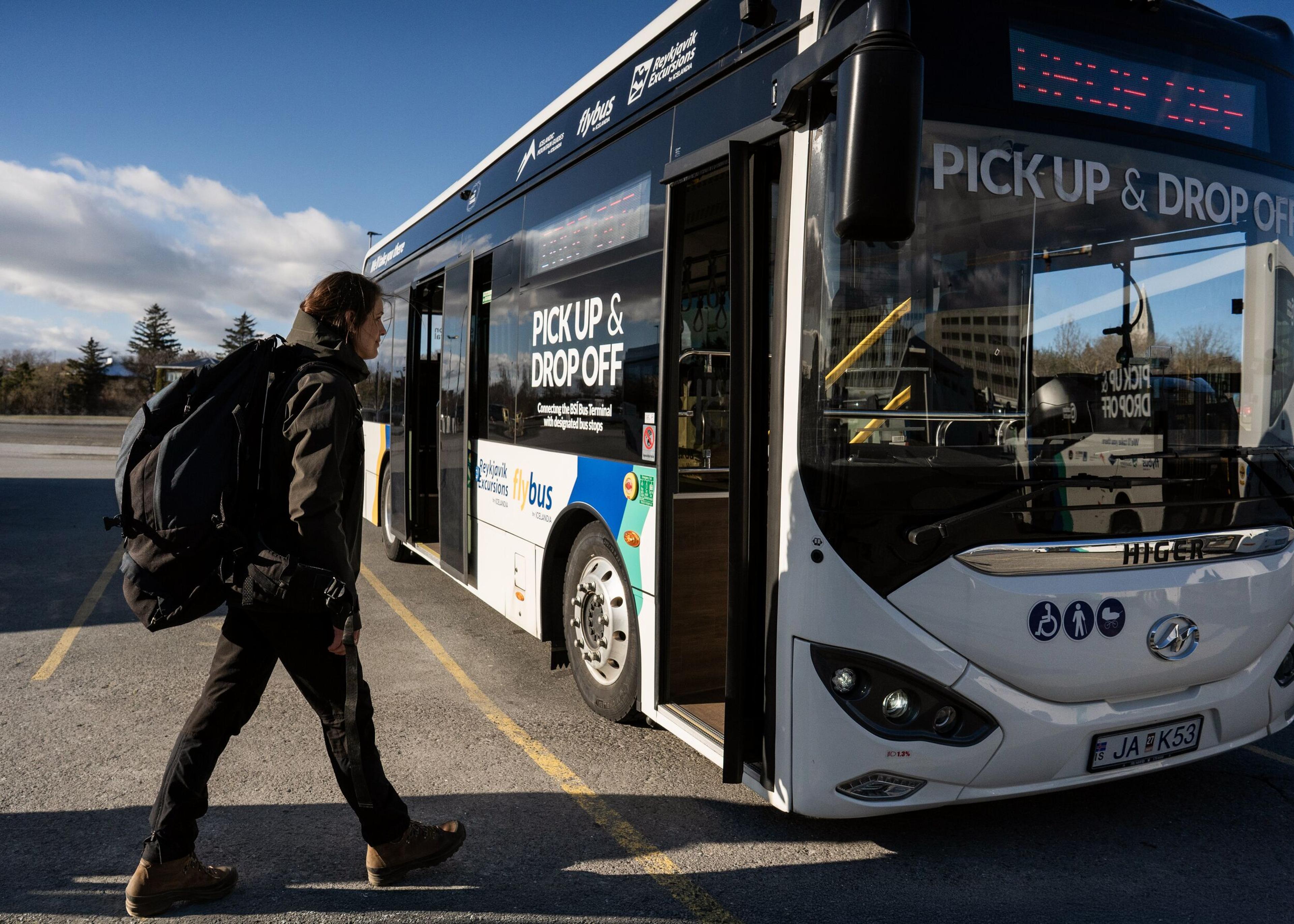 A person with a large backpack walks toward the open door of a white bus labeled "PICK UP & DROP OFF."