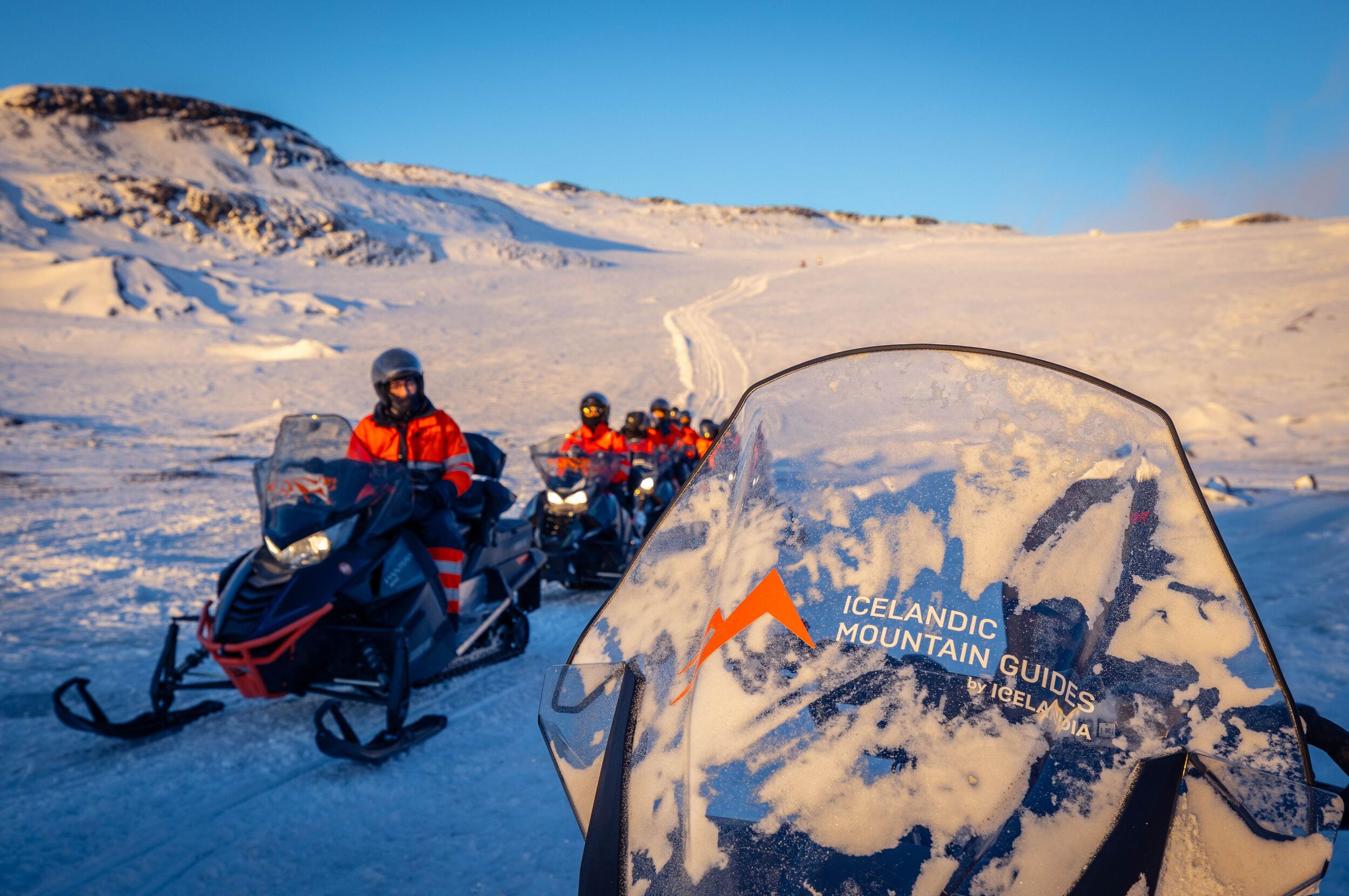 A group of people dressed in orange winter gear rides snowmobiles across a snowy landscape under a clear blue sky.