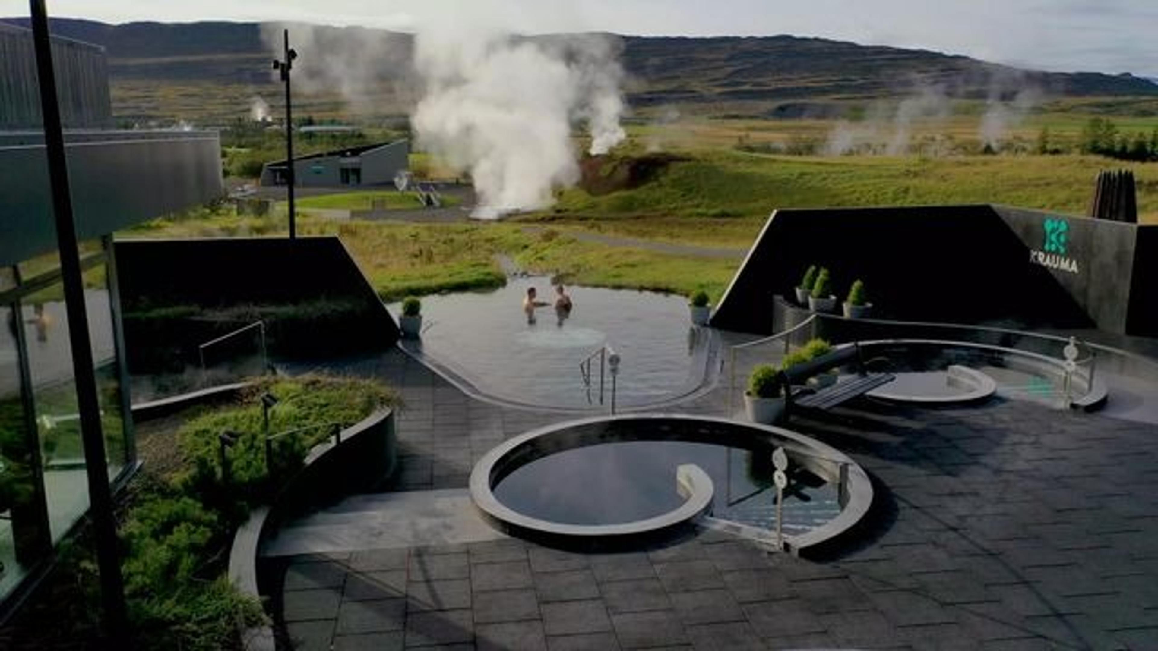 The Krauma geothermal baths in Iceland, featuring circular hot tubs surrounded by modern black stone design and a backdrop of steaming geothermal vents and grassy hills