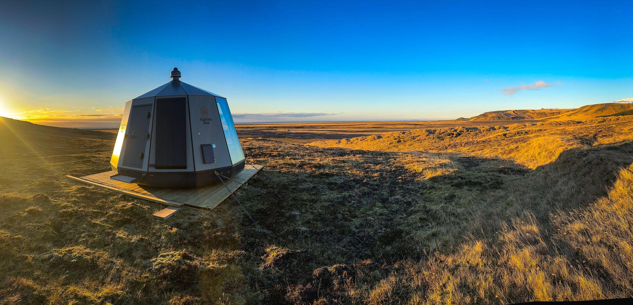 A modern, dark polygonal cabin with large glass panels on a wooden deck in a vast, golden-lit grassy landscape at sunset.