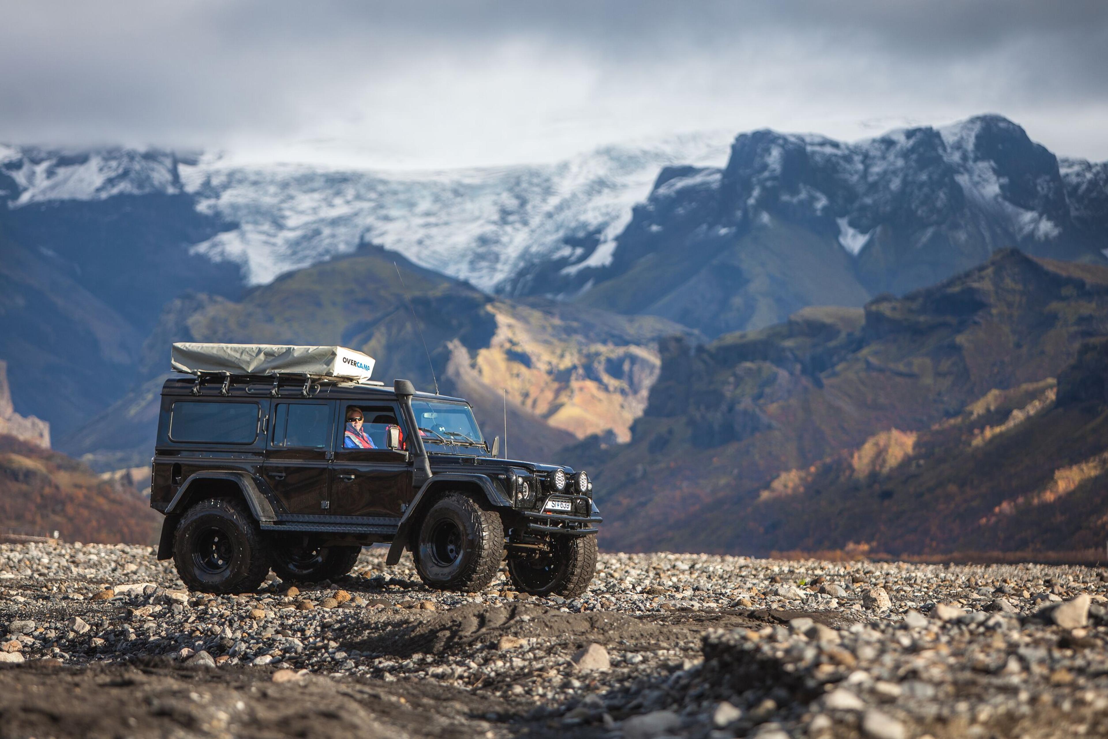 A rugged off-road vehicle equipped with a roof rack travels across a rocky landscape with colorful autumn hills and snow-capped mountains in the background.
