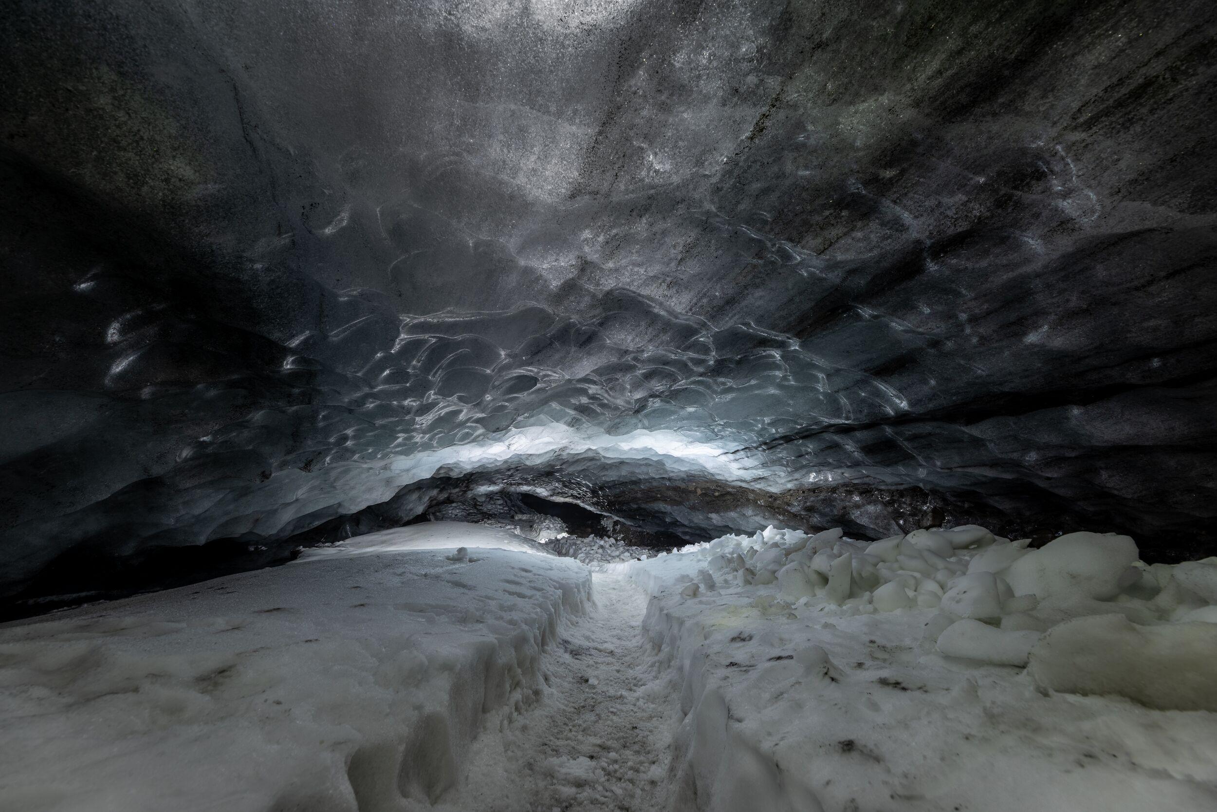 Empty pathway inside Askur ice cave, with textured ice ceiling and snow-covered ground.