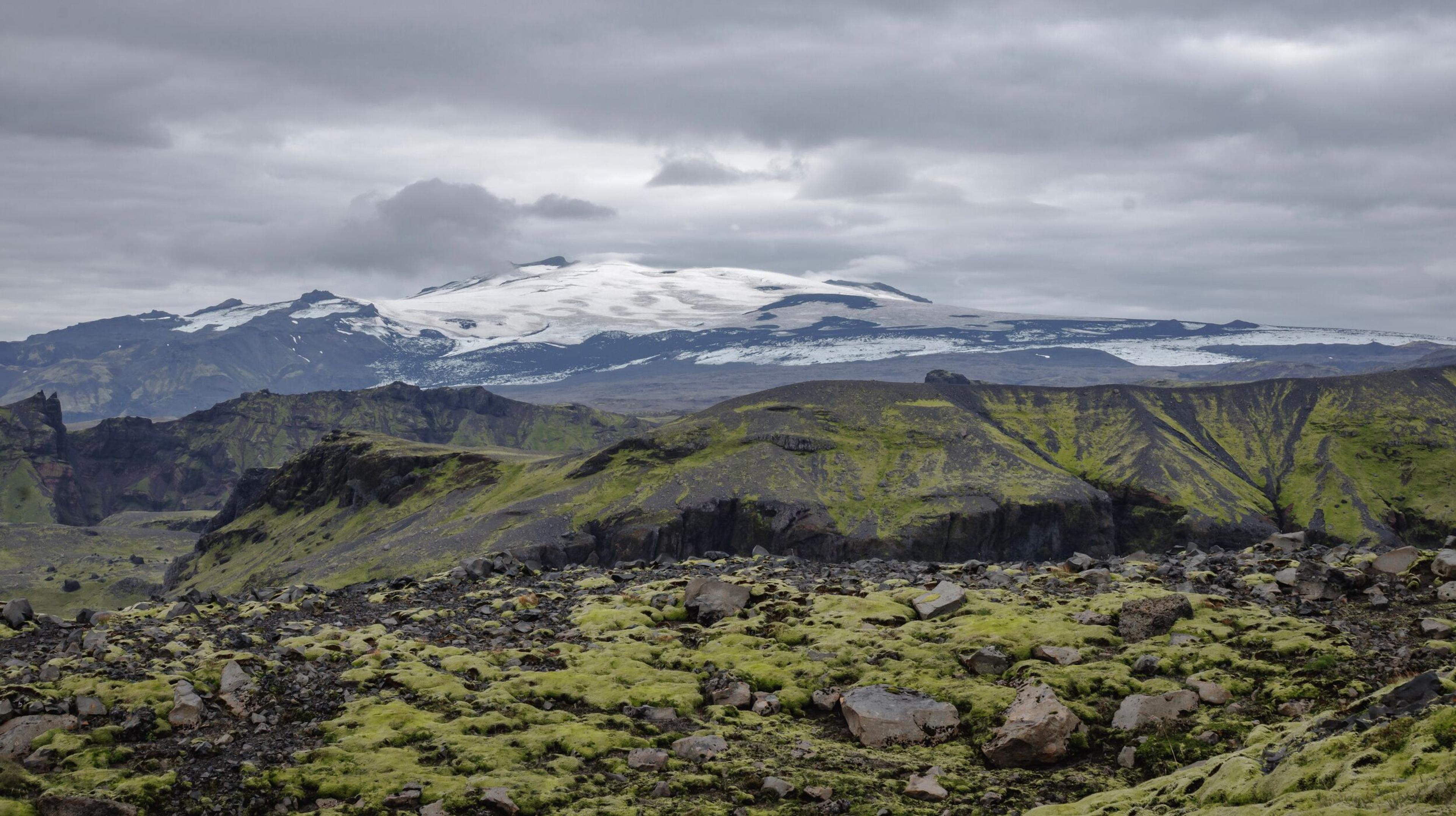 Rocky, moss-covered foreground with dark green hills leading to distant snow-capped mountains under a cloudy sky.