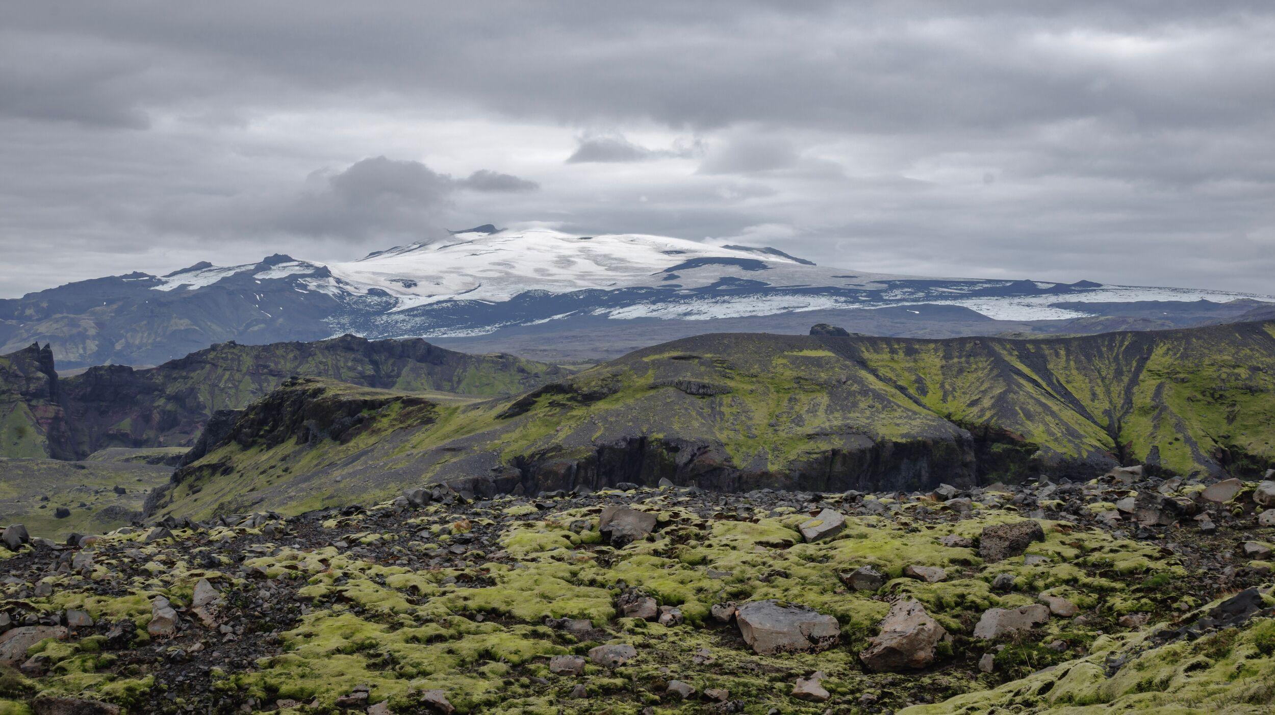 Rocky, moss-covered foreground with dark green hills leading to distant snow-capped mountains under a cloudy sky.
