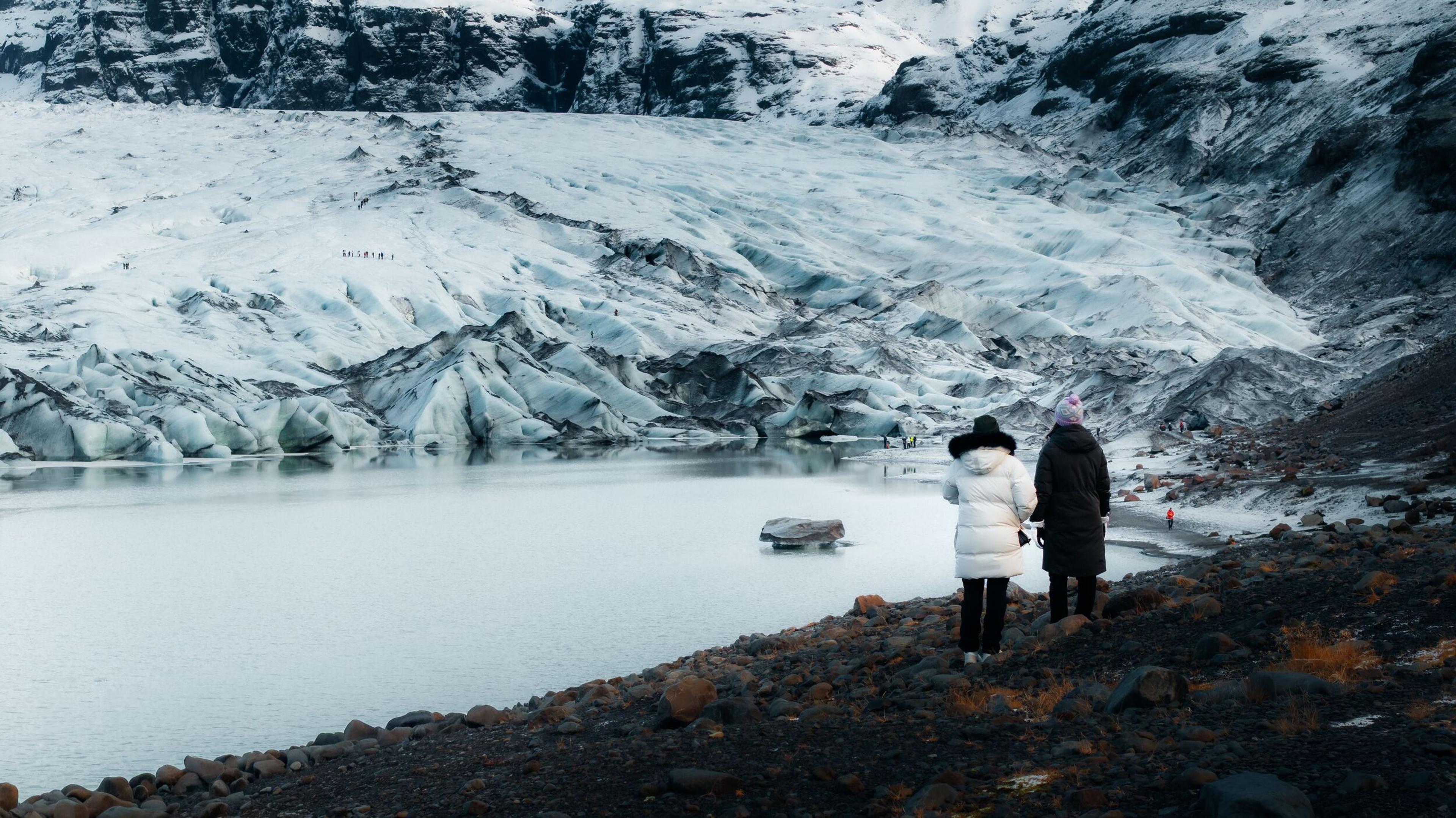 Two people view a sprawling glacier and a tranquil lake from a rocky shore.