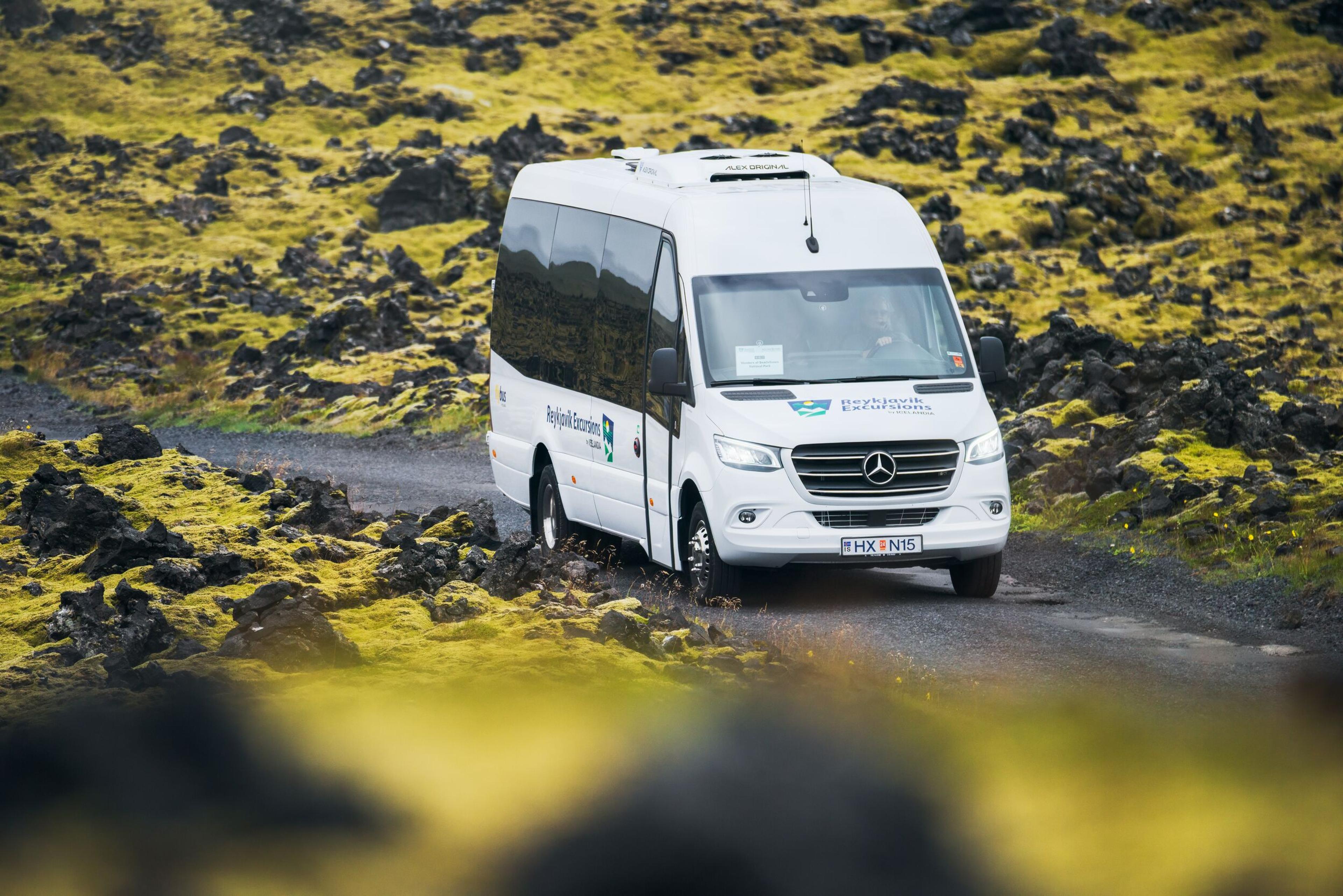 White minibus journeying along a road, surrounded by an expansive moss-covered lava field.