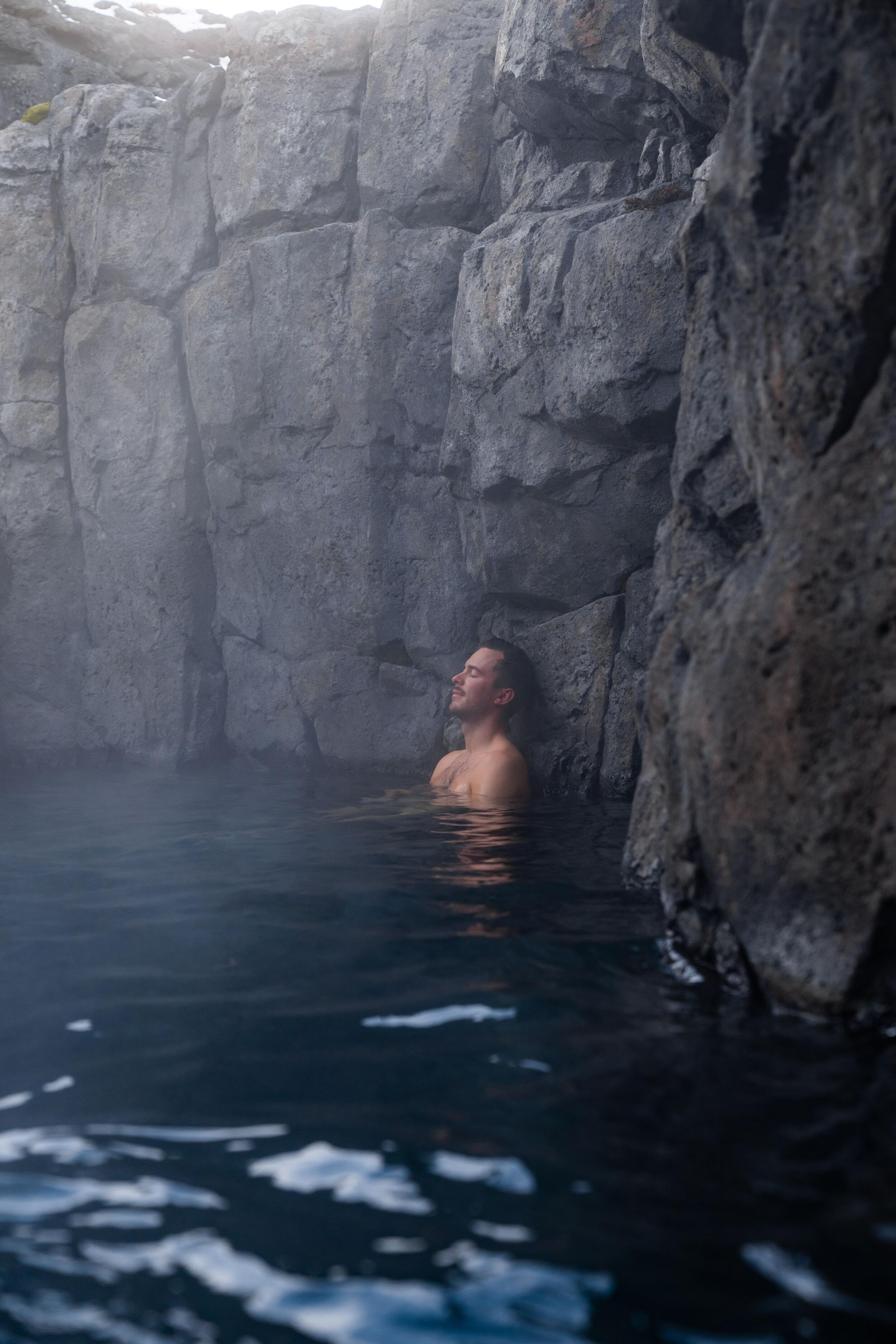 A man leans back against a rocky cliff in the steamy waters of Sky Lagoon, lost in a moment of relaxation and solitude.