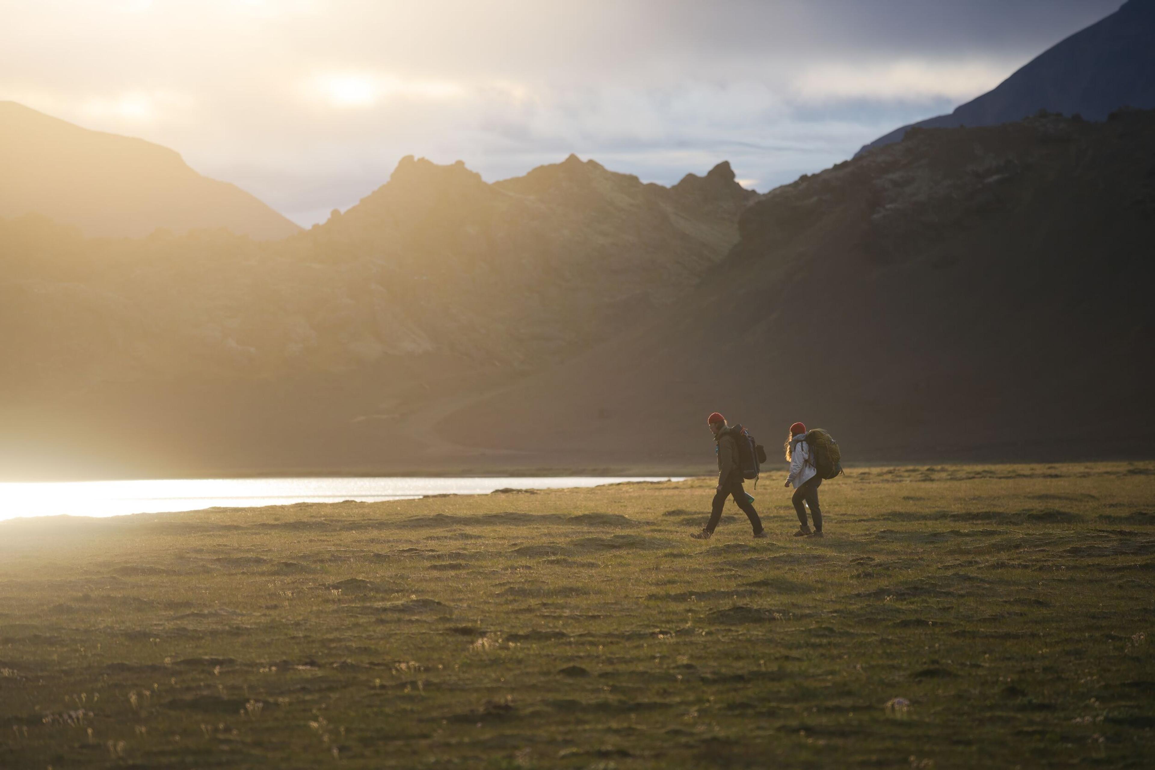 Two hikers with backpacks traverse a grassy field in Iceland, with majestic mountains under the soft glow of sunrise in the background.